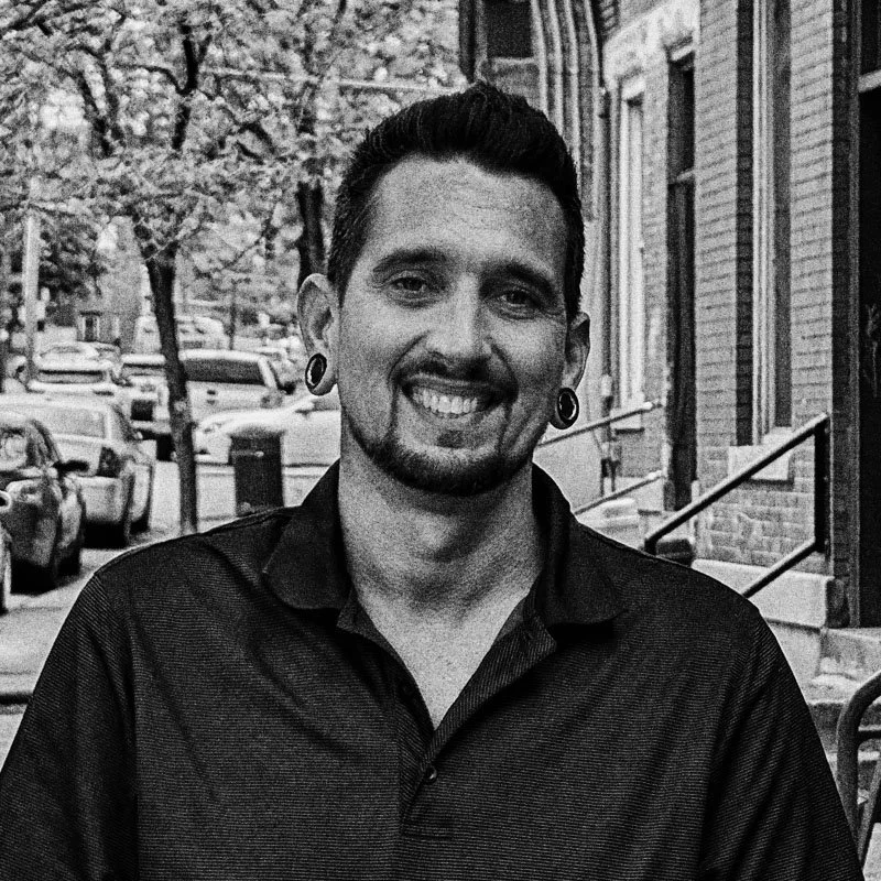 A smiling man with dark hair, gauges in his ears, and a goatee, standing on a city street with cars and trees in the background.