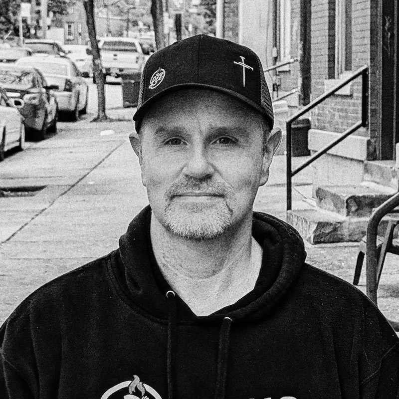 A man with a beard and light-colored hair wearing a black hoodie and a black baseball cap with a cross and a logo. He is sitting outdoors on a city sidewalk with parked cars, stairs, and buildings visible in the background.