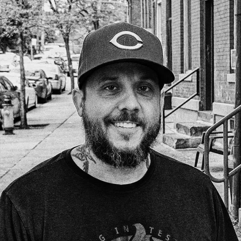 A man with a beard and mustache wearing a Cincinnati Reds baseball cap, smiling, standing on a city sidewalk with cars parked and brick buildings in the background.