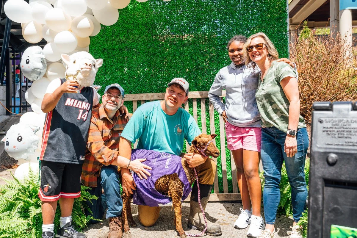 Five people stand around a goat wearing a purple jersey who just won the race. They're smiling and proud.