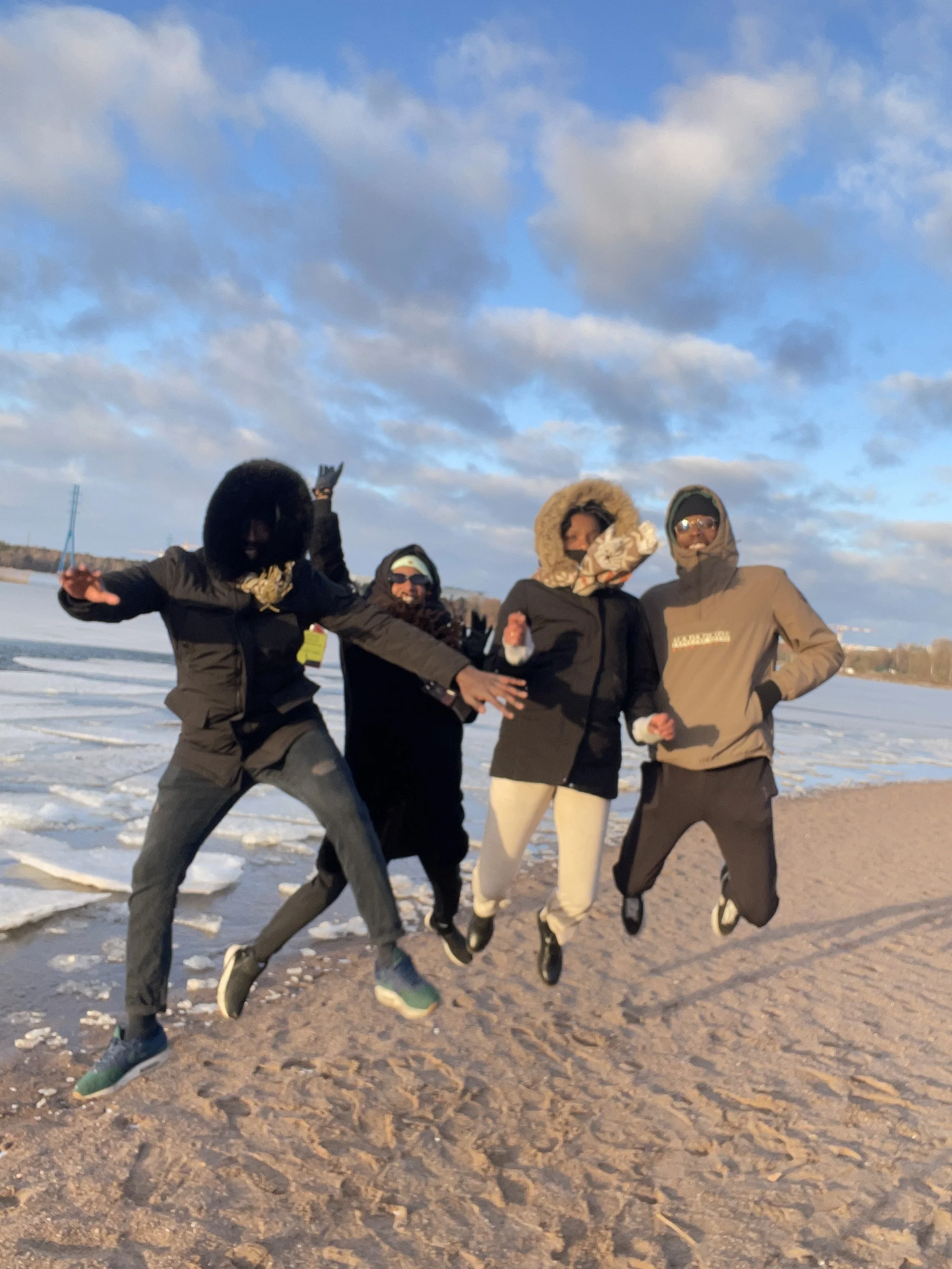 Quatre personnes habillées chaudement sautent sur la plage en hiver, avec le ciel nuageux en arrière-plan.