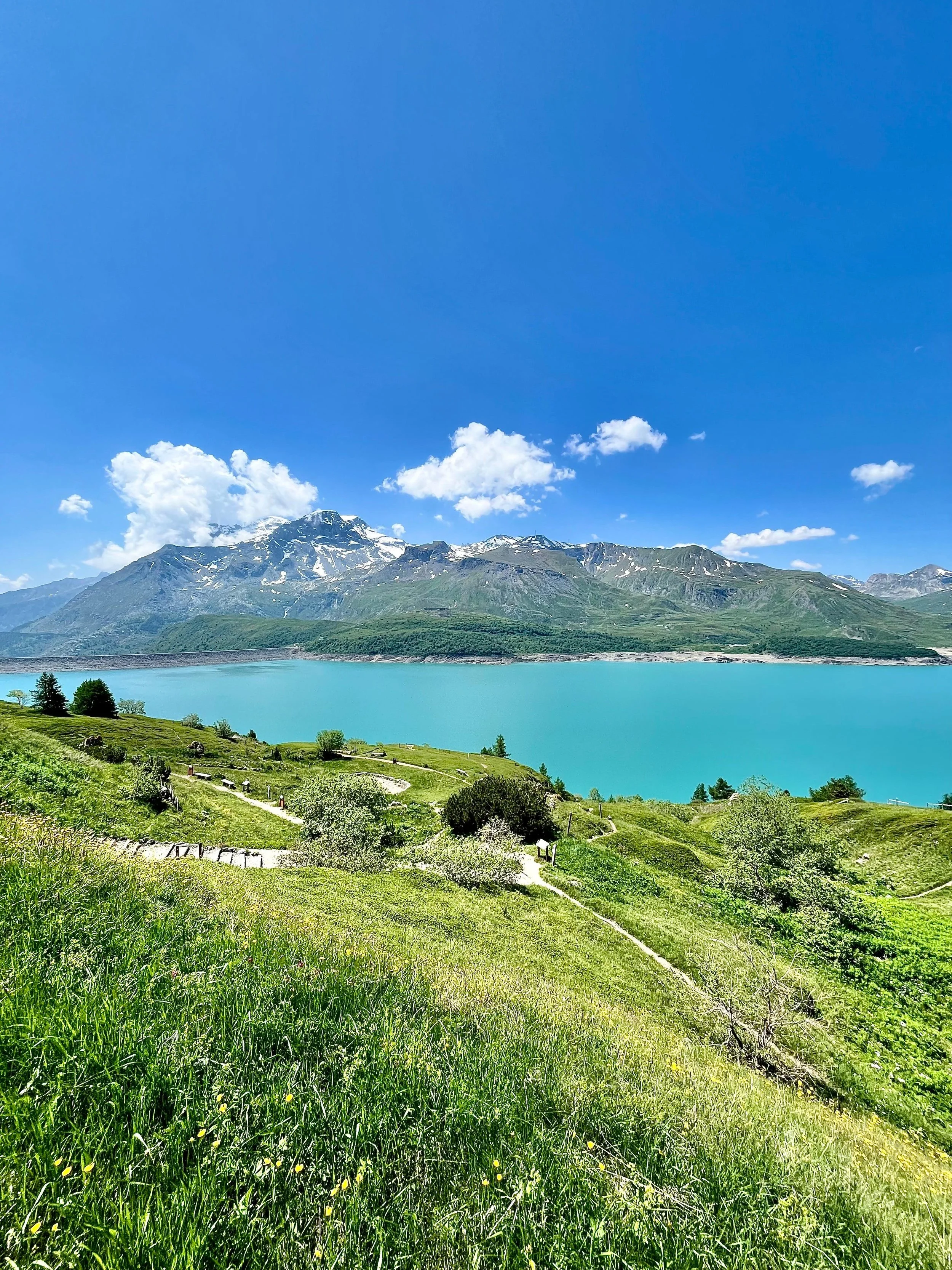 Paysage avec montagnes, lac turquoise, ciel bleu avec quelques nuages, et terrain verdoyant avec des sentiers et des arbres.