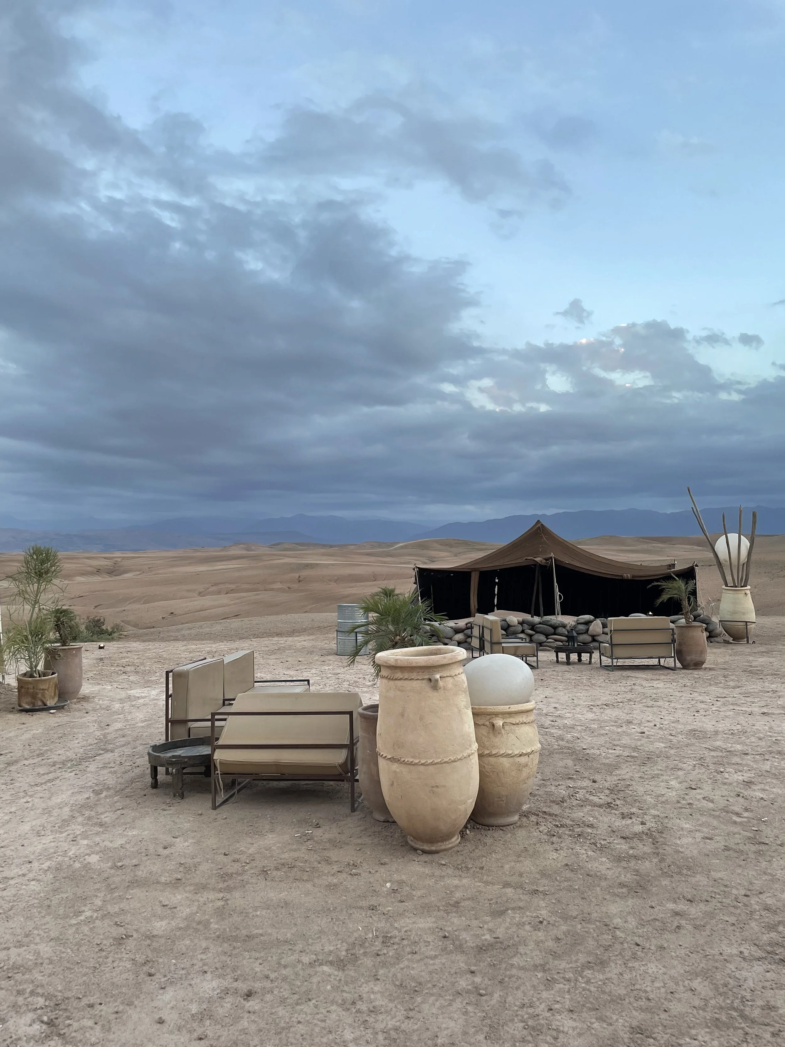 Un campement dans le désert avec une tente en toile, des meubles de salon, des pots en terre cuite et des plantes décoratives, sous un ciel nuageux, avec des montagnes à l'horizon.
