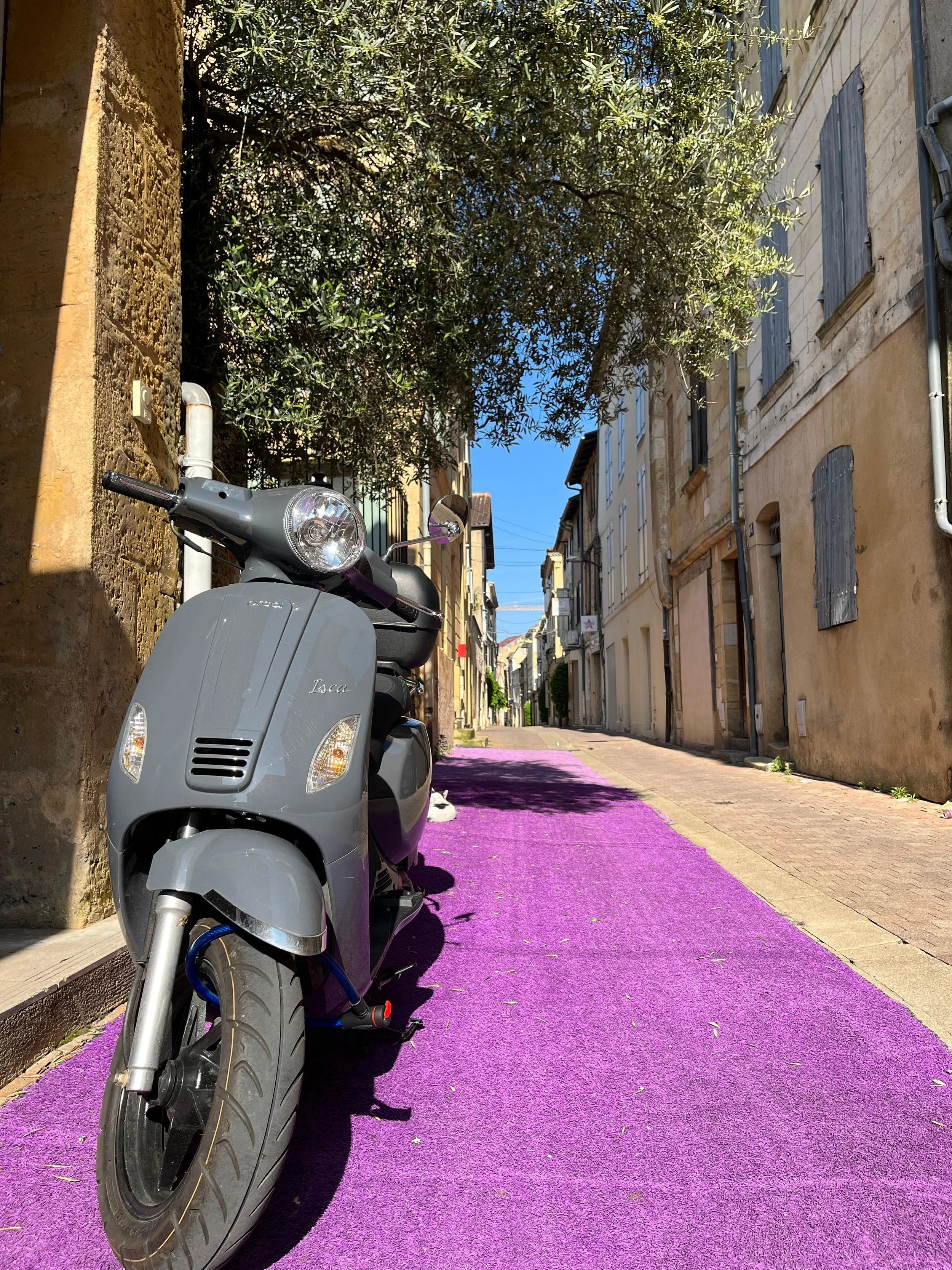 Scooter gris stationné sur une voie pavée violette dans une rue étroite avec des bâtiments en pierre et des fenêtres à volets en bois, ombragée par un arbre.