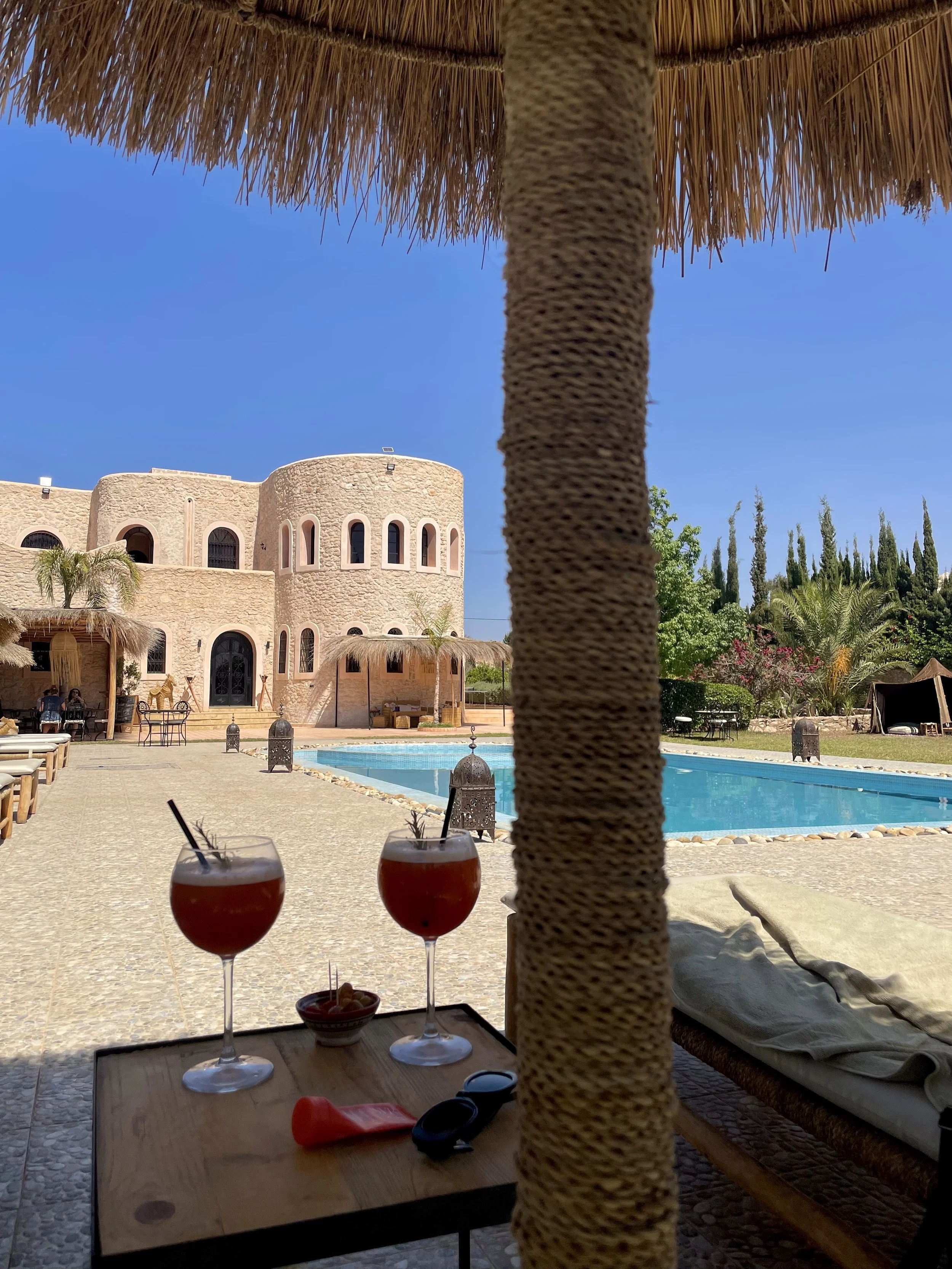 Une vue d'une piscine dans une résidence de style méditerranéen avec un bâtiment en pierre, des palmiers, un lit de soleil et deux cocktails rouges avec des glaçons, sous un parasol en paille.
