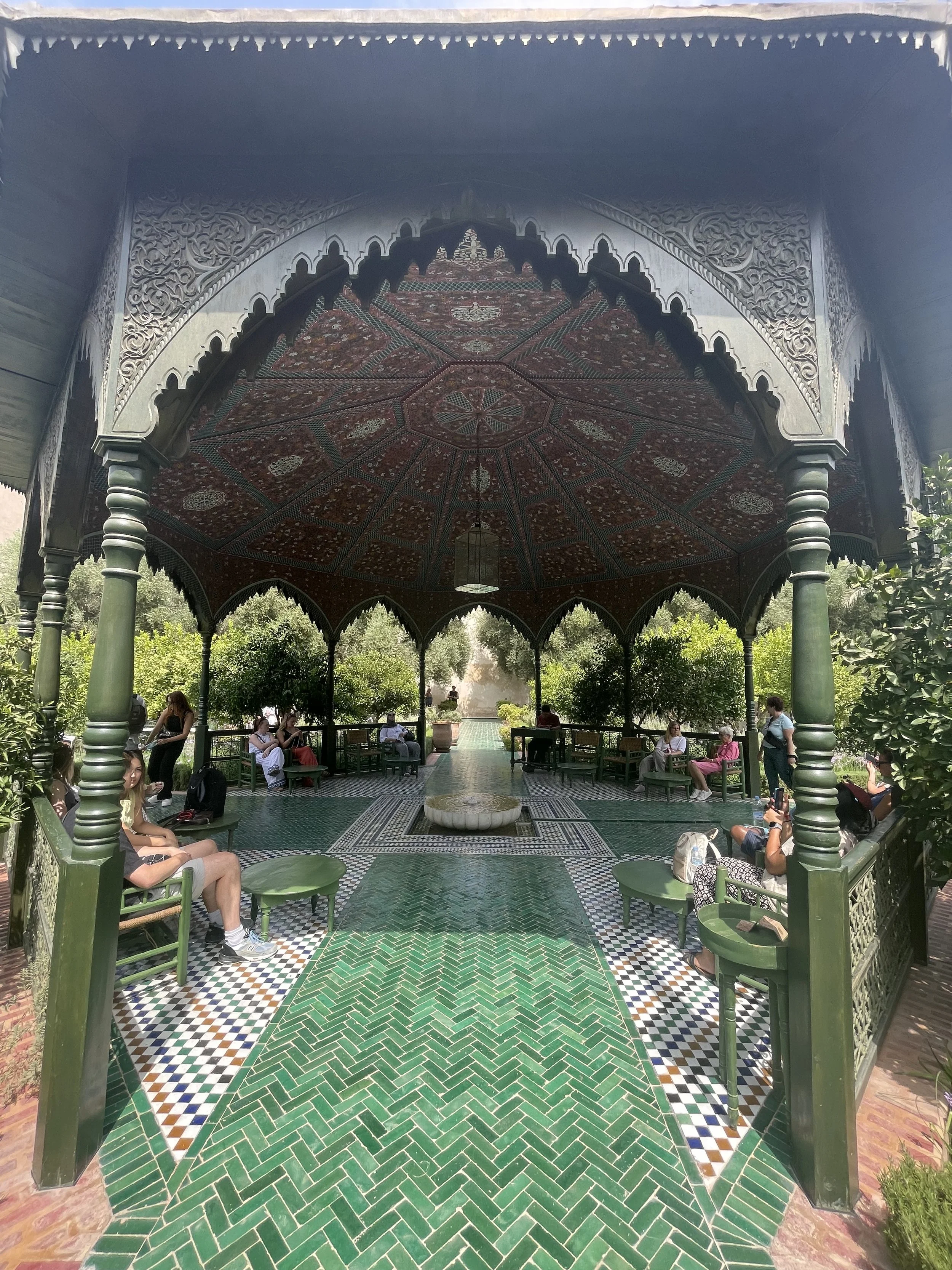 Pavillon marocain traditionnel avec mosaïque, colonnes vertes, plafond orné, personnes assises et se détendant dans un jardin.