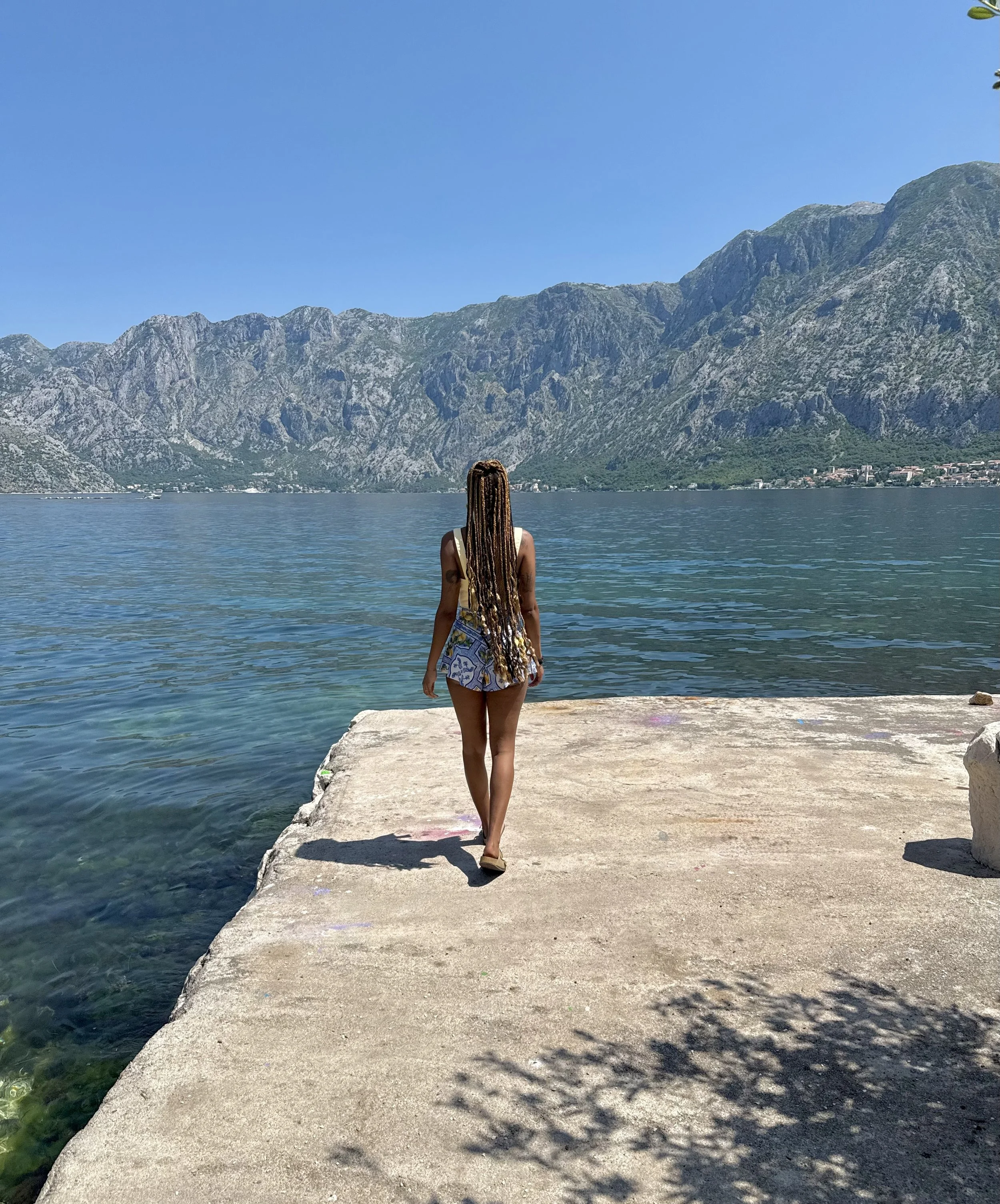 Une femme avec de longs cheveux tressés marche vers la mer depuis un quai en pierre, avec des montagnes en arrière-plan et un ciel bleu clair.