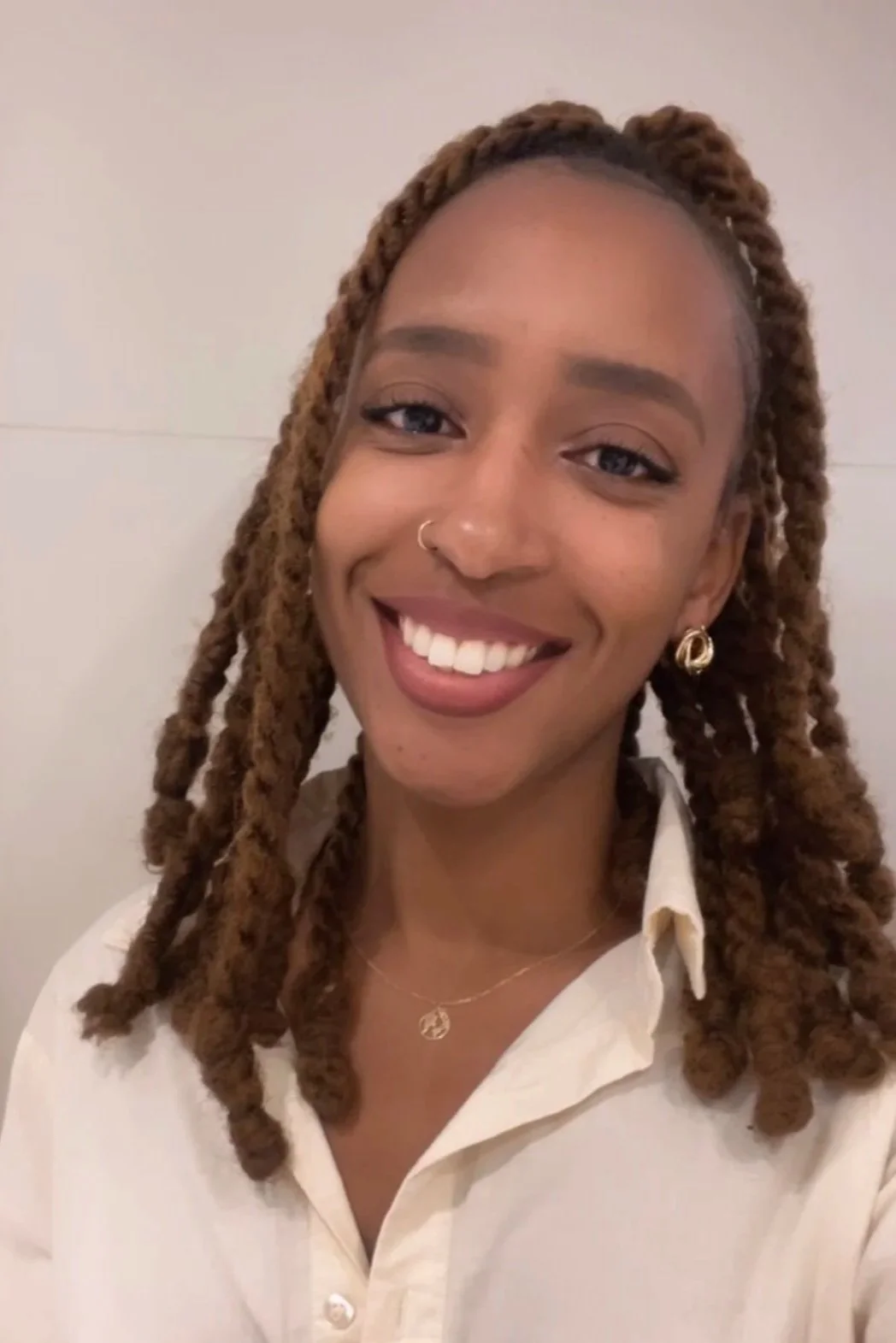 Une jeune femme souriante avec des dreadlocks bruns, portant une chemise blanche, des boucles d'oreilles en or, un collier et un piercings au nez, contre un fond neutre.