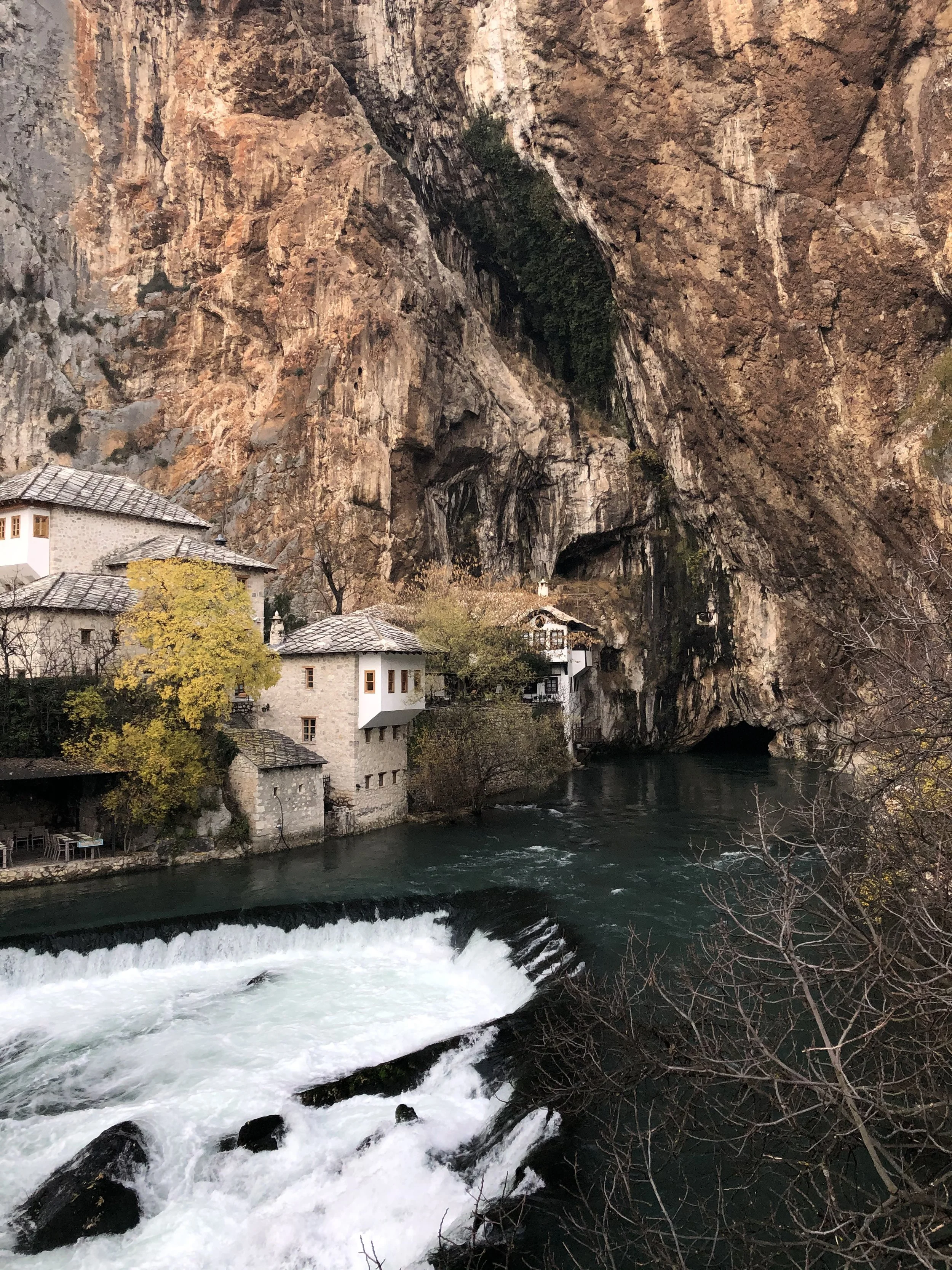 Maisons en pierre traditionnelles au bord d'une rivière avec une cascade, entourées de falaises rocheuses.