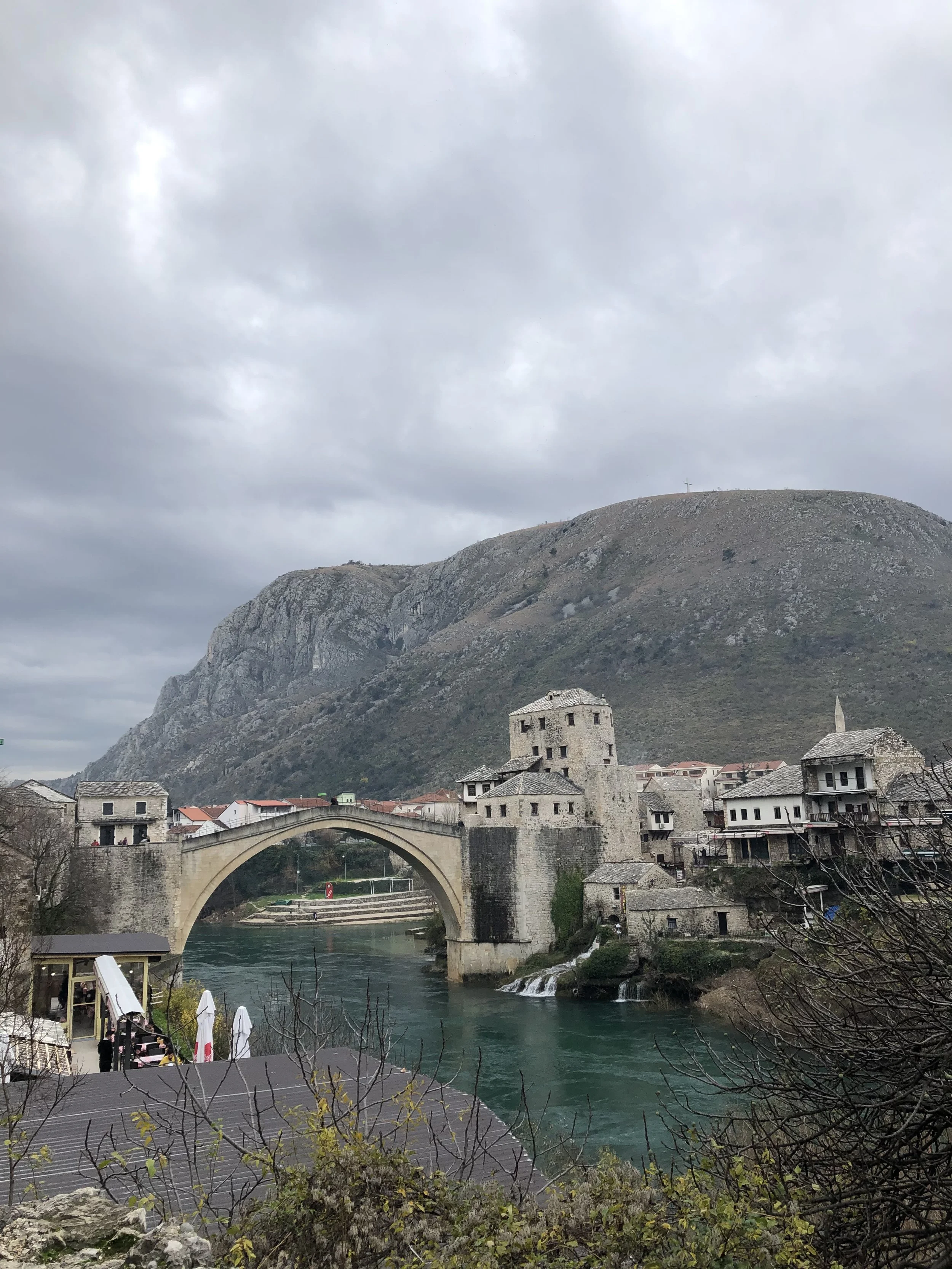 Paysage urbain avec un pont en pierre traversant une rivière turquoise, un château en pierre et une vieille ville avec des montagnes en arrière-plan, sous un ciel nuageux.