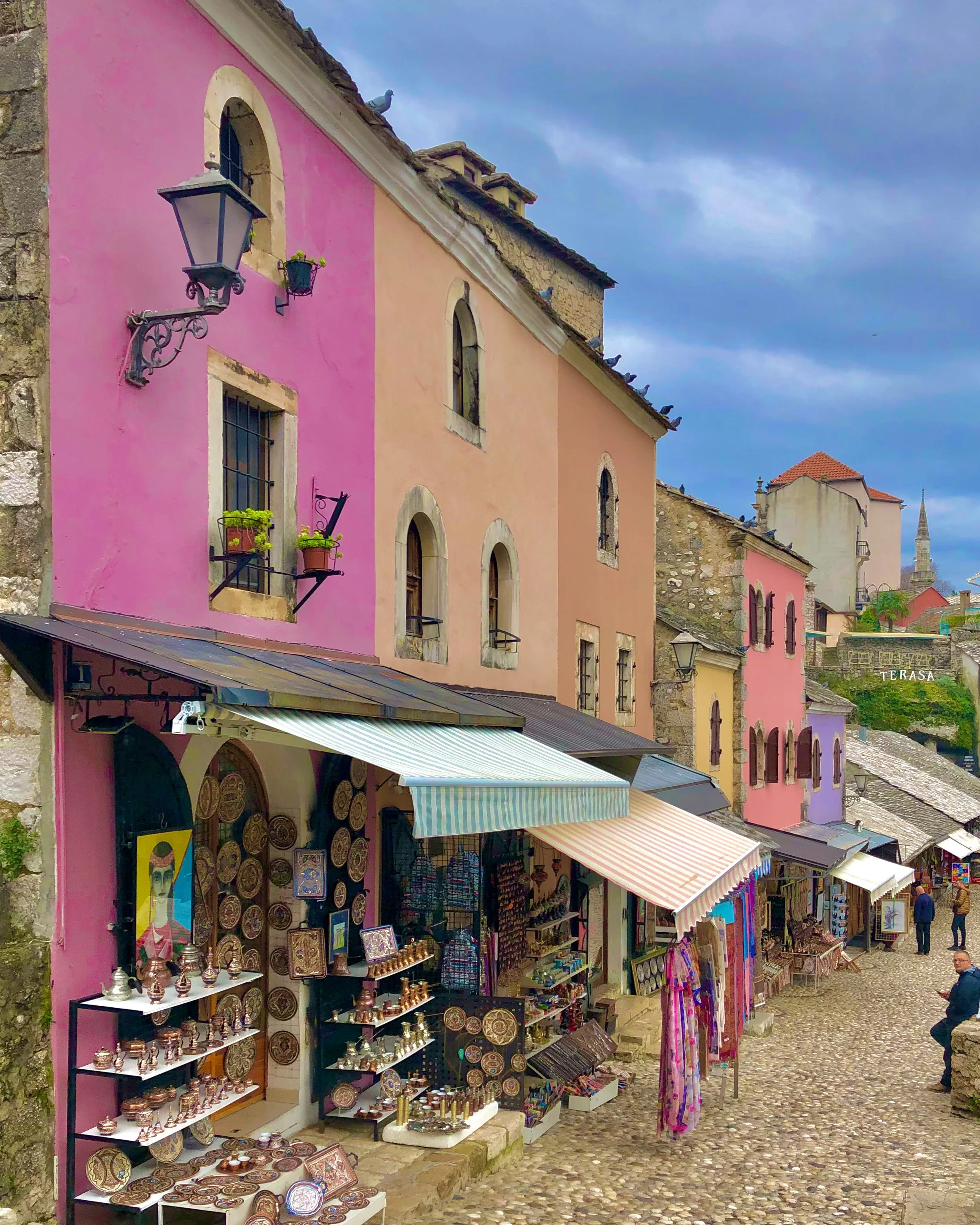 Rue pavée avec des boutiques colorées, façades en rose et pastel, étals d'artisanat, et quelques passants, dans un village pittoresque.