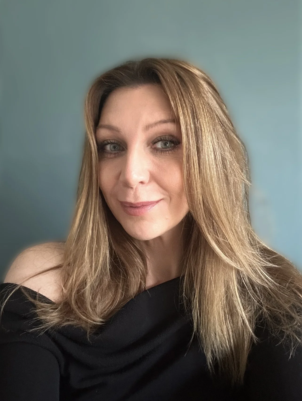 A woman with long, wavy, light brown hair and blue eyes smiling softly, wearing a black off-shoulder top against a teal background.