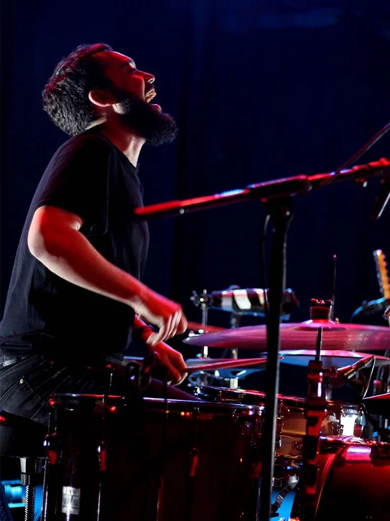 Concerts. A man with a beard and dark hair playing the drums on stage, illuminated by colorful stage lights.