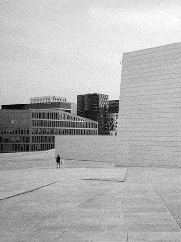 Analog Black and white photo of modern buildings with a solitary person walking on a wide open paved space.