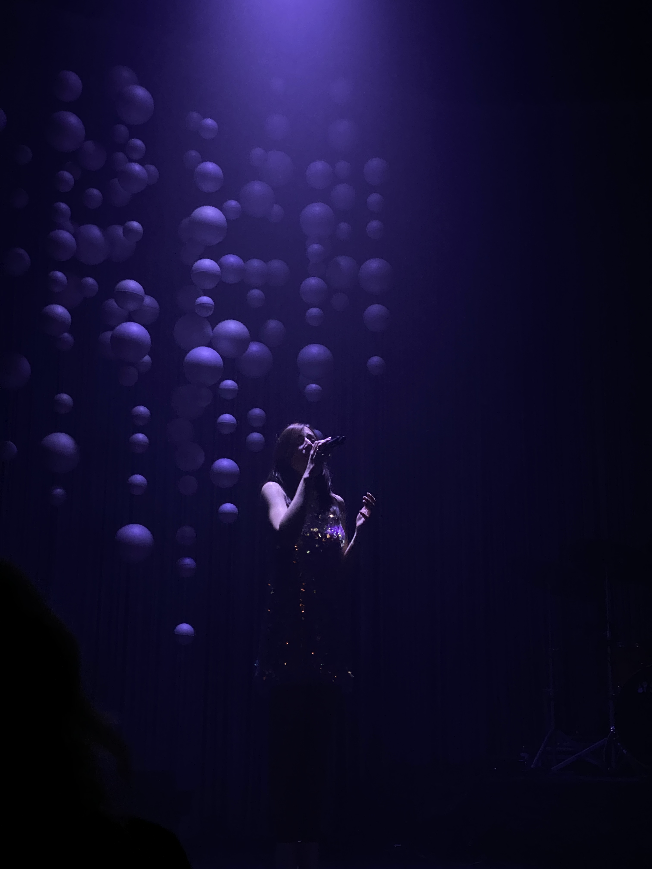 Concerts. songweiter CARLA. A woman singing on a stage with hanging purple balls as decoration, illuminated by purple stage lighting.