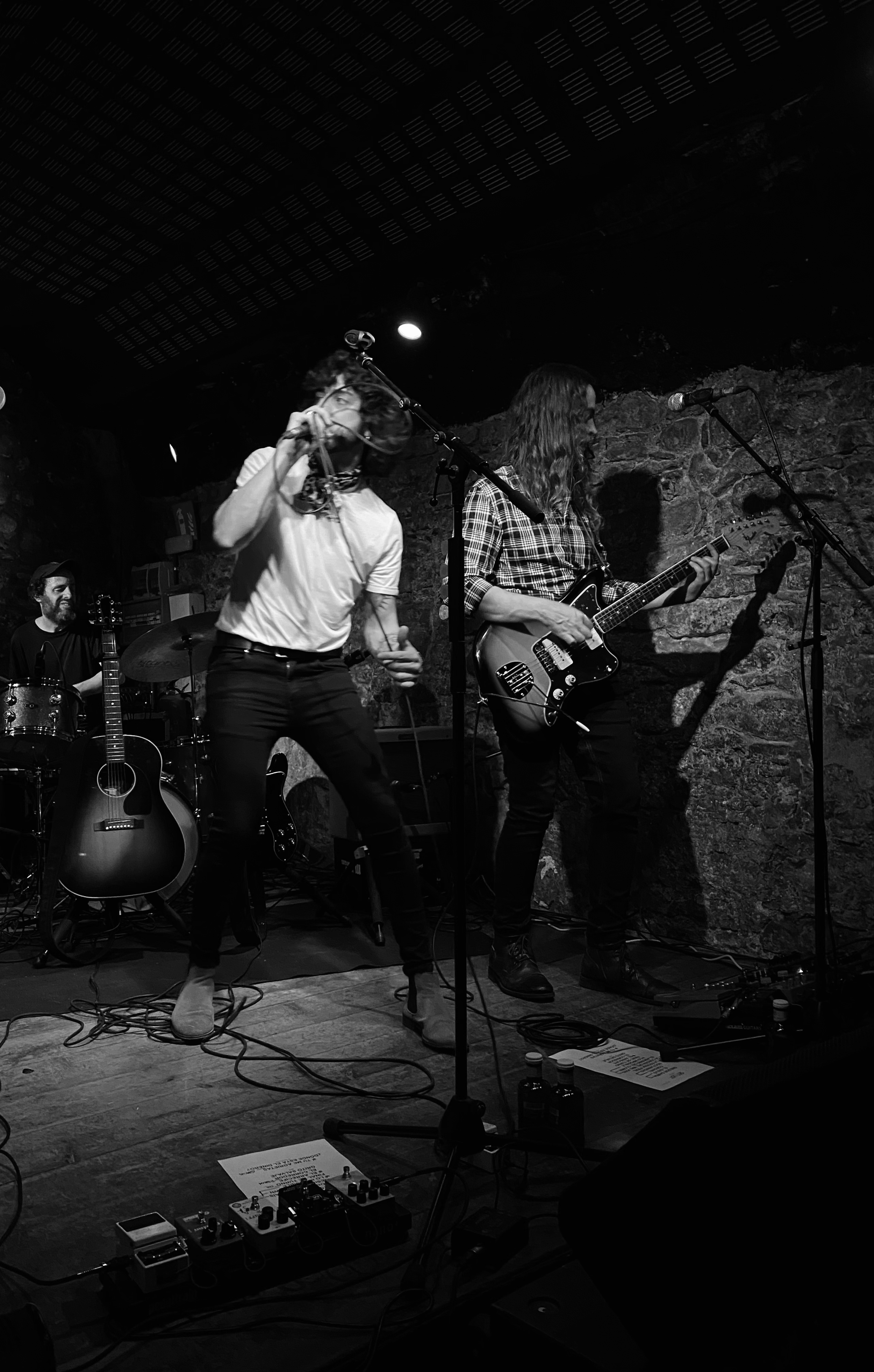 Concerts. Songwriter Daniel Boga. Musicians performing on stage at a bar or club, including a man singing and playing drums, and a woman playing electric guitar, with a brick wall background and music equipment on stage.