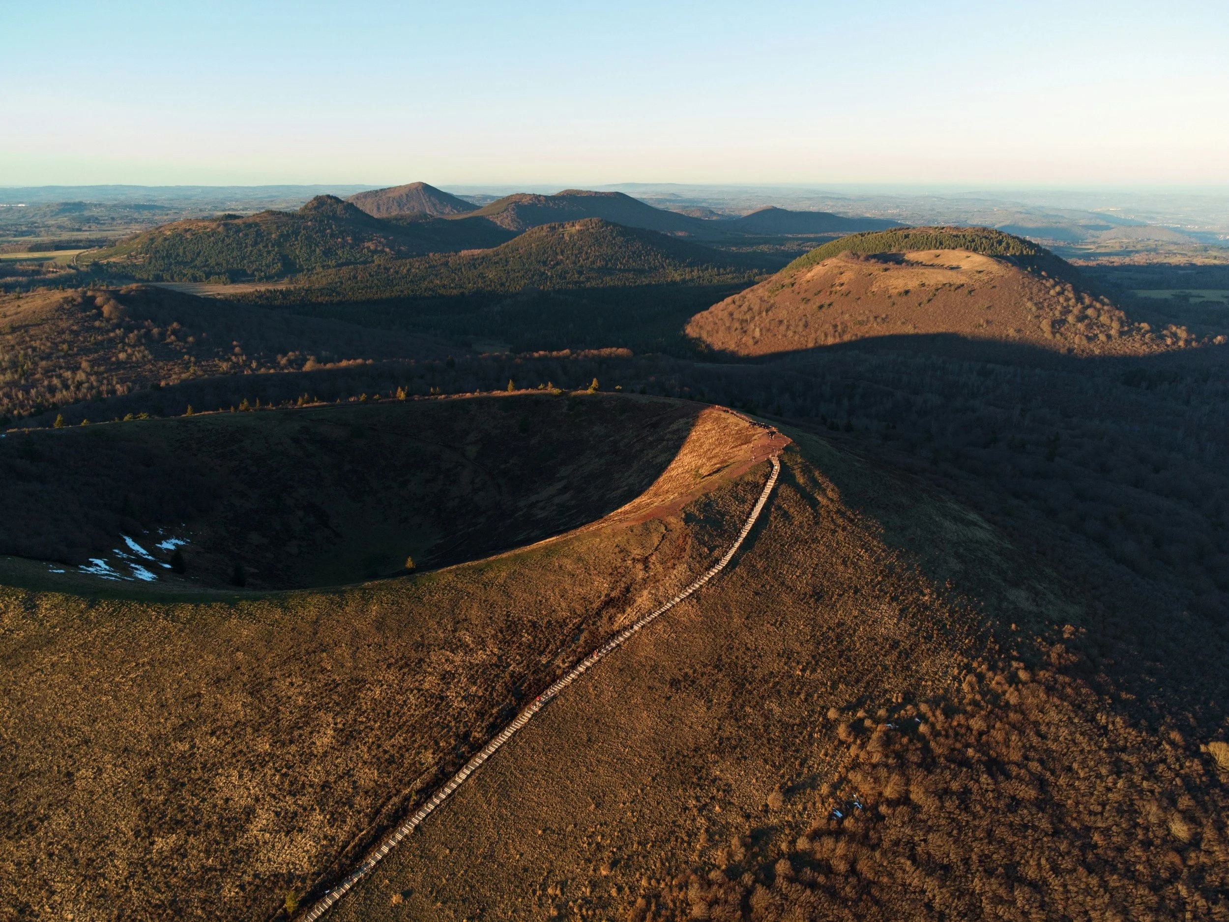 Vue aérienne de collines et montagnes avec un sentier serpentant sur la crête d'une colline, en partie éclairée par le soleil.