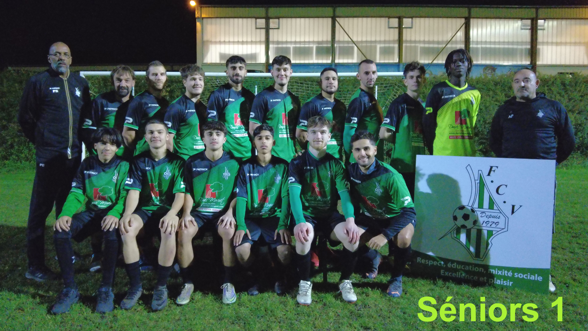 Une équipe de football composée de joueurs et d'entraîneurs posant pour une photo sur un terrain en dehors, avec un panneau indiquant F.C.V. et la mention "Depuis 1979". Il s'agit d'une équipe de seniors 1. La photo est prise la nuit avec un éclairage artificiel.