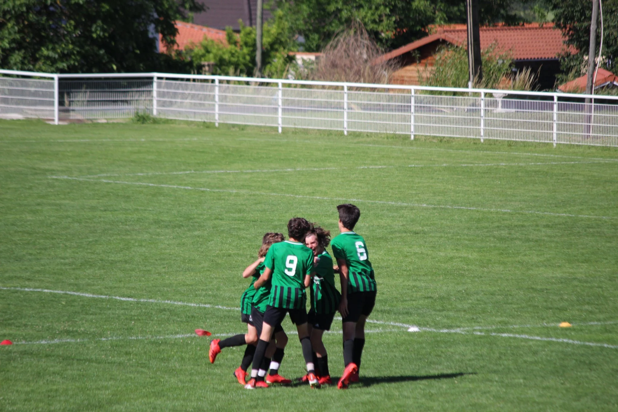 Groupe de jeunes joueurs de football en vert célèbrent une victoire sur un terrain en herbe avec une clôture blanche en arrière-plan.