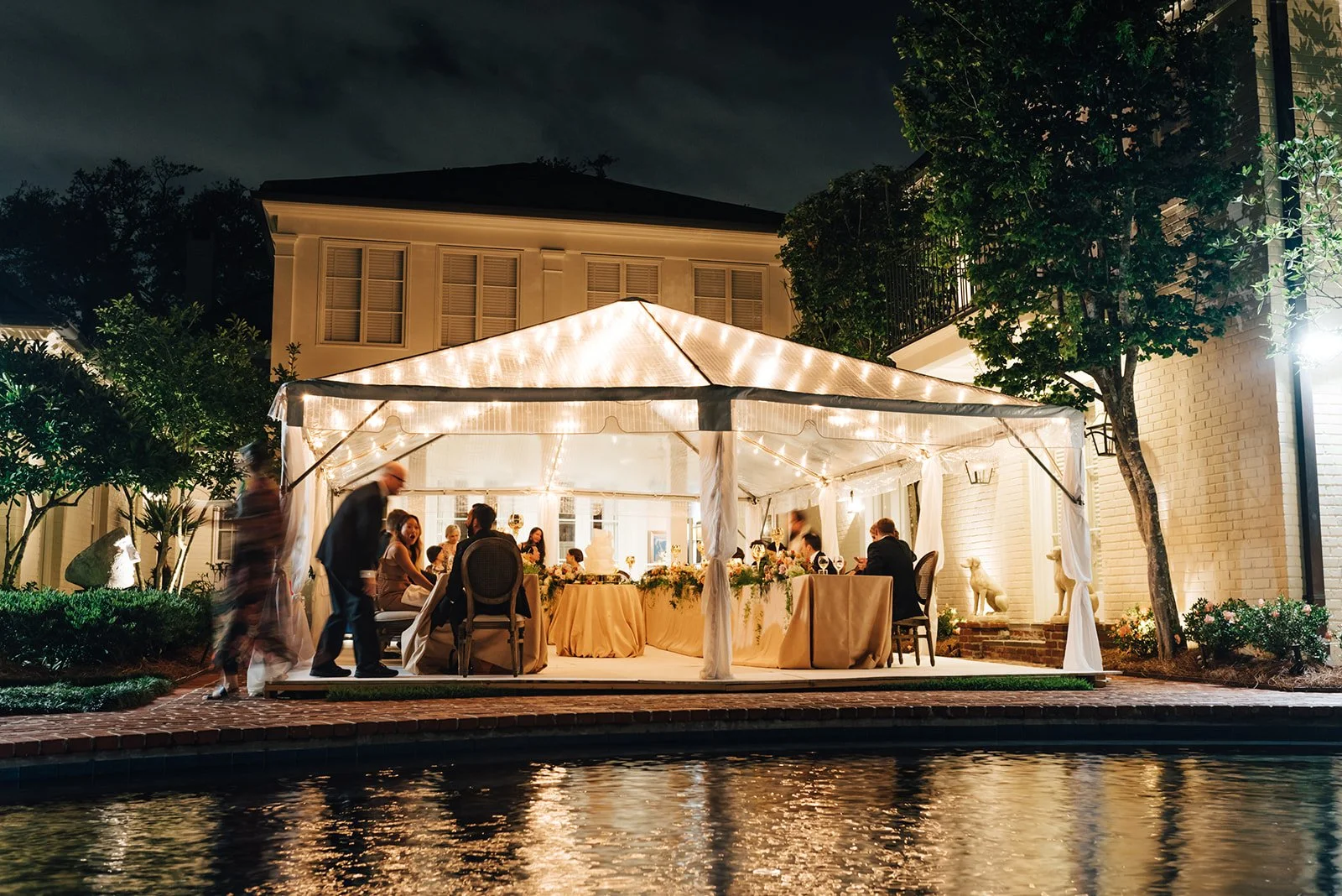 Nighttime outdoor gathering under a decorated white canopy tent illuminated with string lights, with people sitting at tables and socializing, near a swimming pool with reflections of the lights on the water, surrounded by trees and a house.