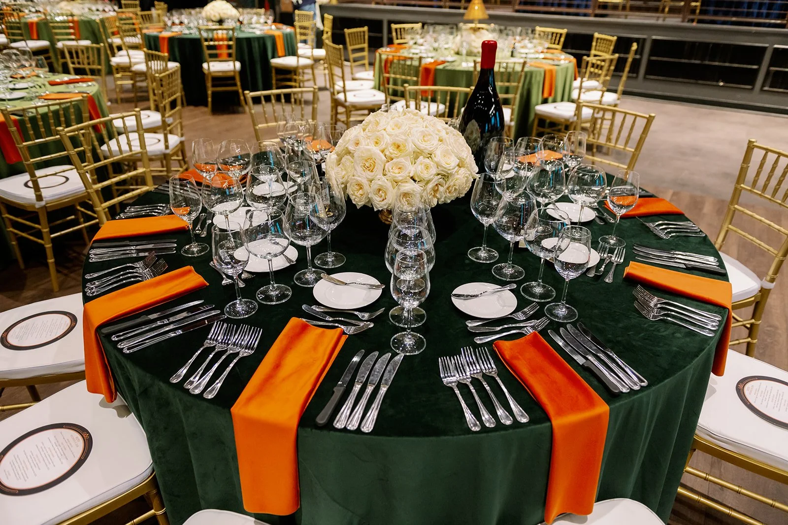 A round banquet table set for a formal event with multiple wine glasses, plates, silverware, and orange napkins, decorated with a bouquet of white roses and a large wine bottle in the center, surrounded by gold chairs.