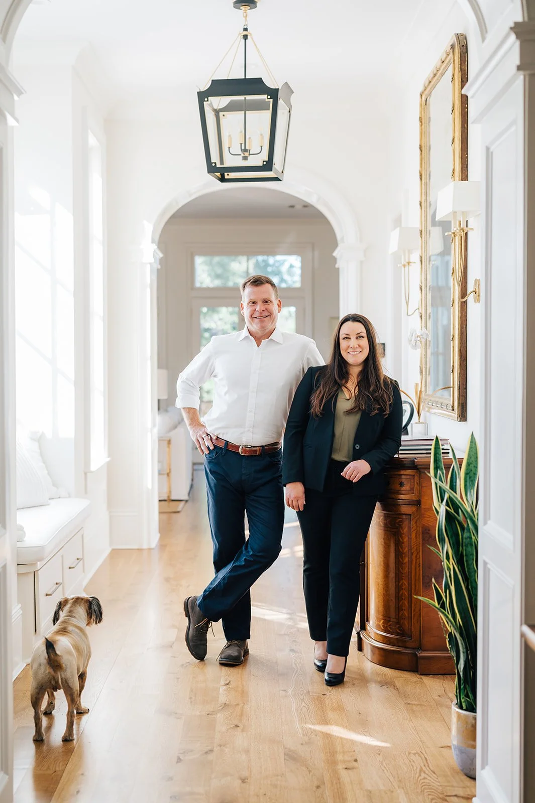 A man and woman standing in a well-lit, elegant hallway of a home, smiling, with a small dog on the floor nearby.