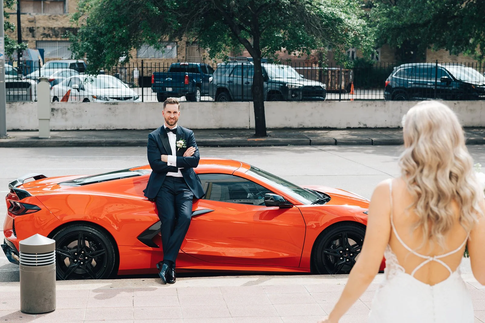 A groom in a navy tuxedo with a white shirt and black bow tie standing with arms crossed next to an orange sports car on the street. A bride with long, curly blonde hair and a white lace wedding dress is approaching from the foreground at a wedding o