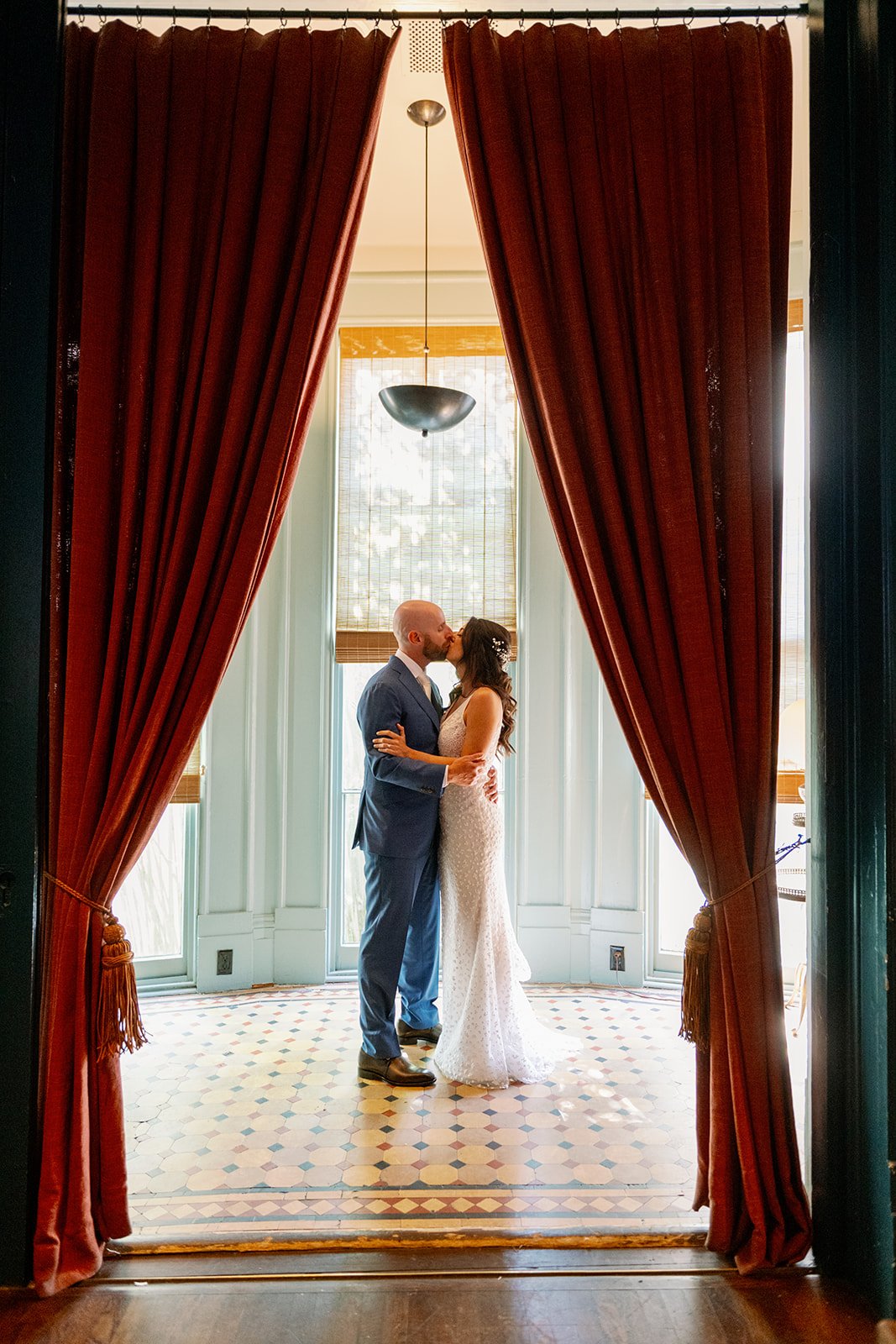 A couple kissing, dressed in wedding attire, standing in front of large windows with natural light, framed by red curtains, with patterned tile floor beneath them.