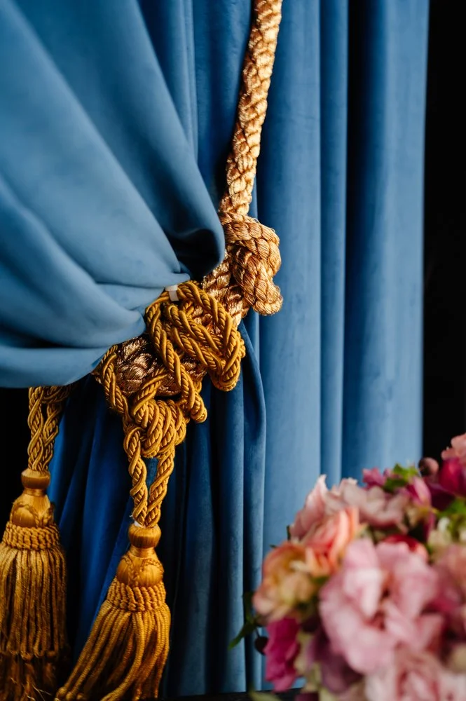Close-up of a blue curtain tied with ornate gold and beige tassel tiebacks, with pink flowers partially visible in the foreground.