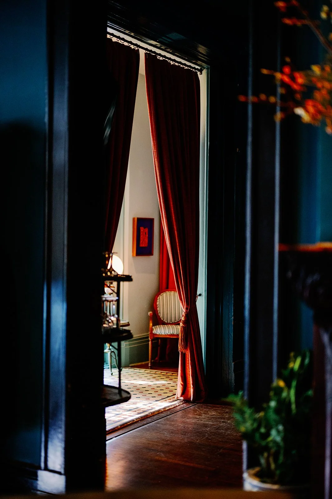 A doorway with red velvet curtains partially drawn, revealing a vintage striped armchair, a side table with a lamp, a framed artwork on the wall, and a patterned tile floor in a cozy room.