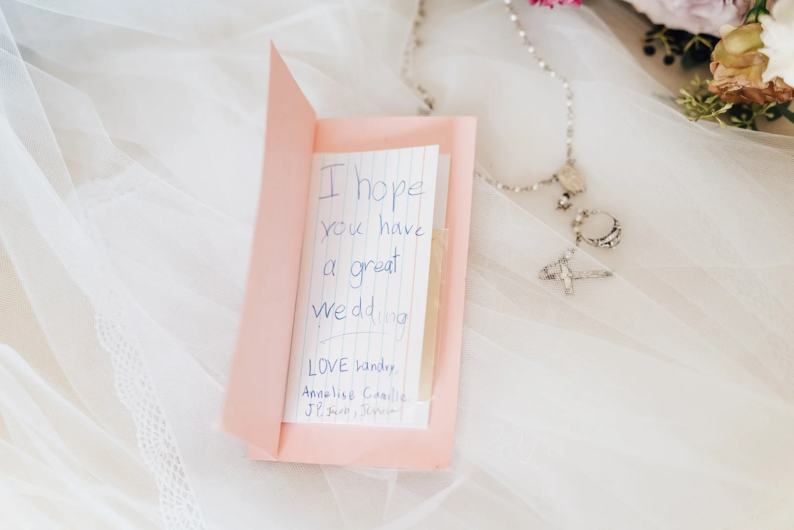 A pink wedding card with handwritten message, placed on white lace fabric. The message reads "I hope you have a great wedding. LOVE Landry, Annelise, Camille, JP, Jacob, Jasmine." Next to the card are silver jewelry pieces including a necklace with a