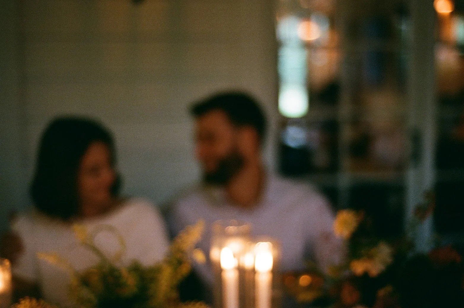 A blurred photo of a man and woman sitting together at a table with candles and flowers, in a dimly lit setting.