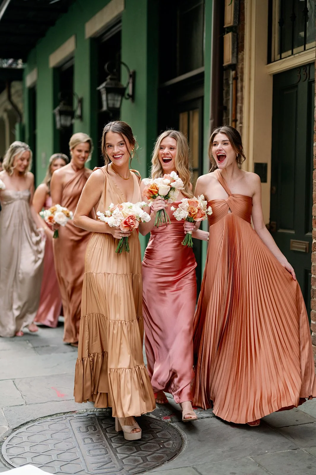 A group of women in satin and pleated dresses, holding bouquets, walking together on a city sidewalk, smiling and laughing.
