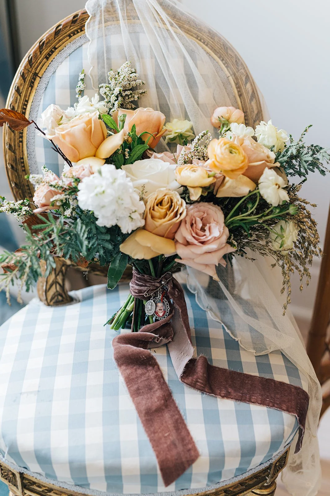 A bouquet of mixed pink and white roses, cream-colored flowers, and greenery on an ornate chair with a blue and white checkered cushion, with tulle fabric and a brown velvet ribbon attached.