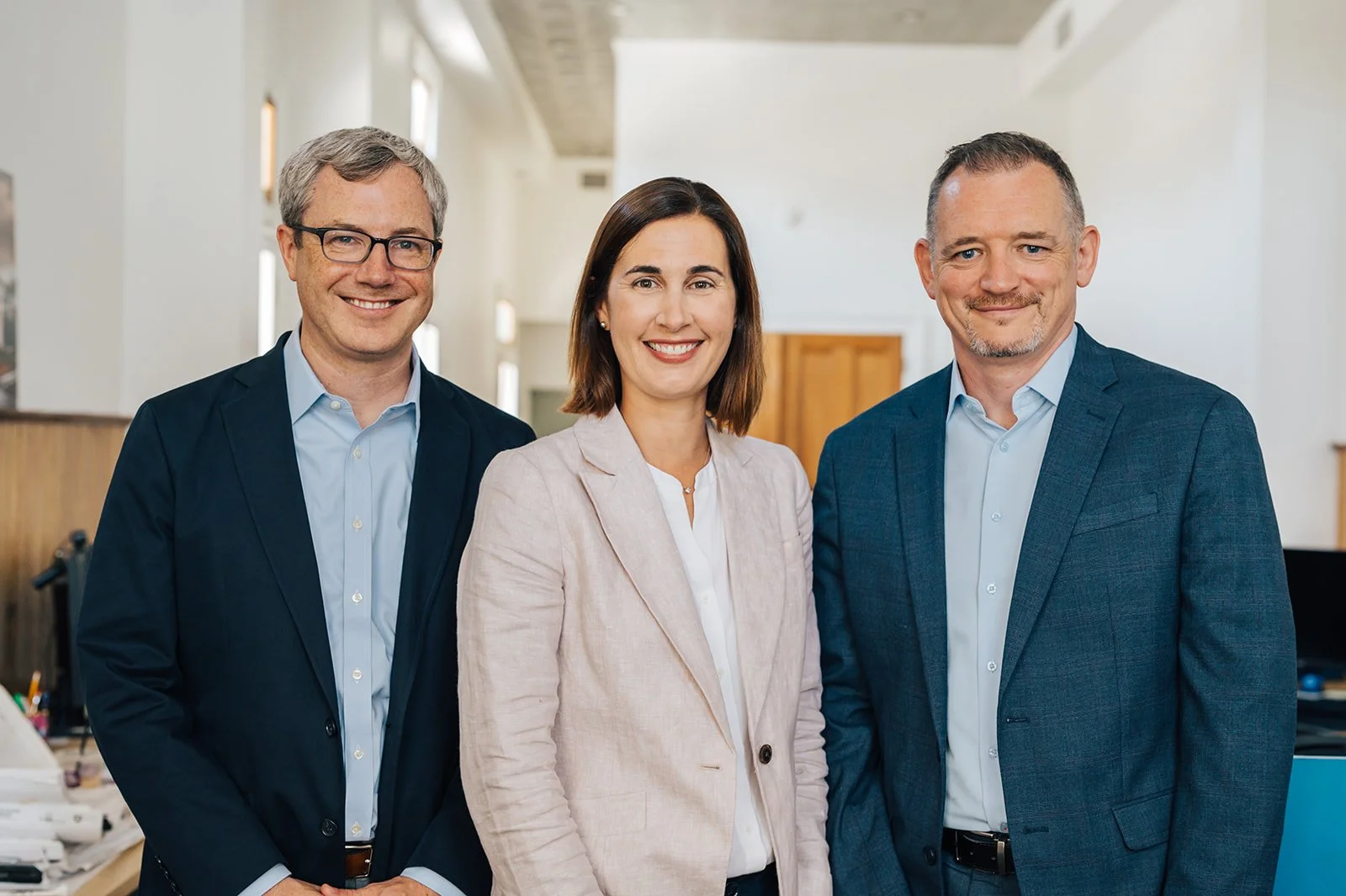 Three professionally dressed people, two men and one woman, smiling and standing indoors in an office or conference room.