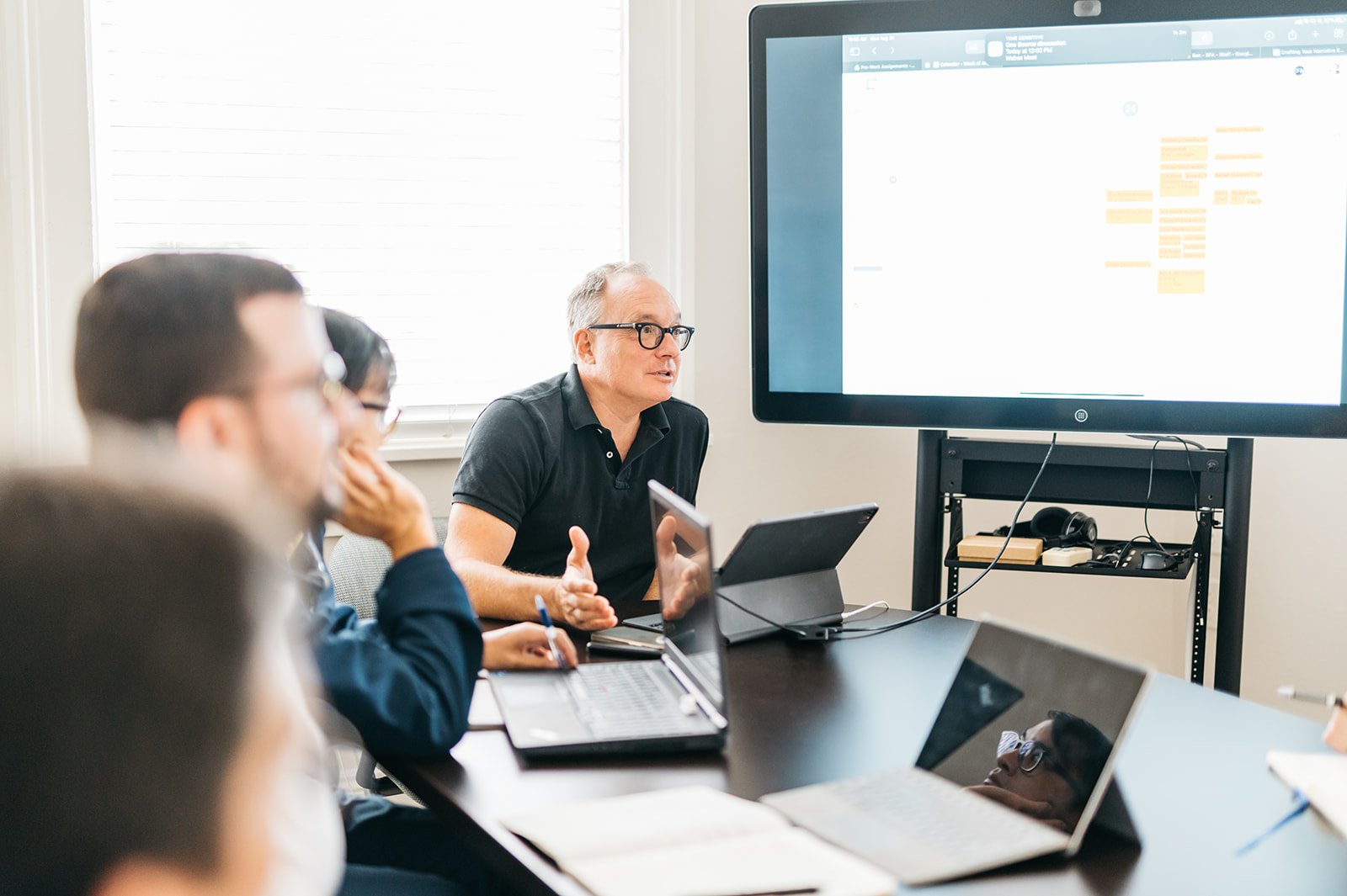 A group of professionals in a meeting room watching a presentation on a large screen, with laptops and notebooks on the table.