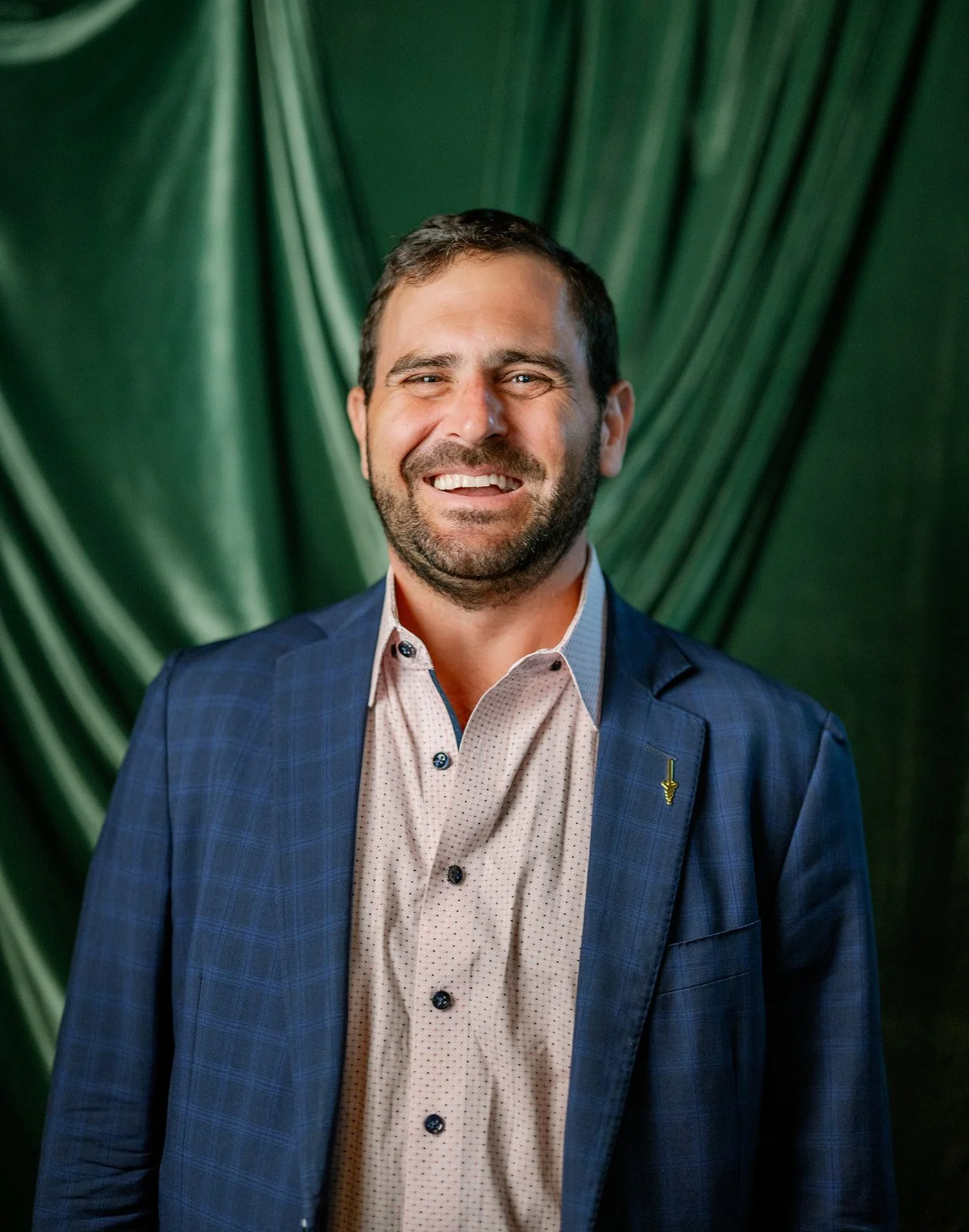 A man with dark hair and a beard wearing a blue suit jacket and a light-colored button-up shirt, smiling, with a green fabric background.