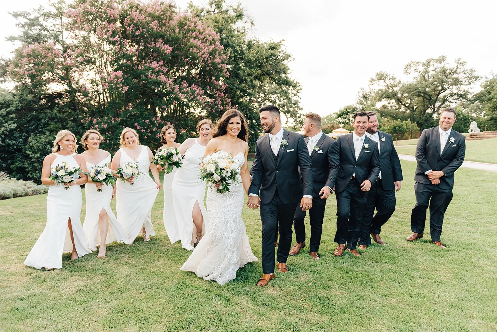 A wedding party walking on a grassy field with women in white dresses holding bouquets and men in dark suits with boutonnières, smiling and enjoying each other's company, with trees and pink flowering bush in the background.
