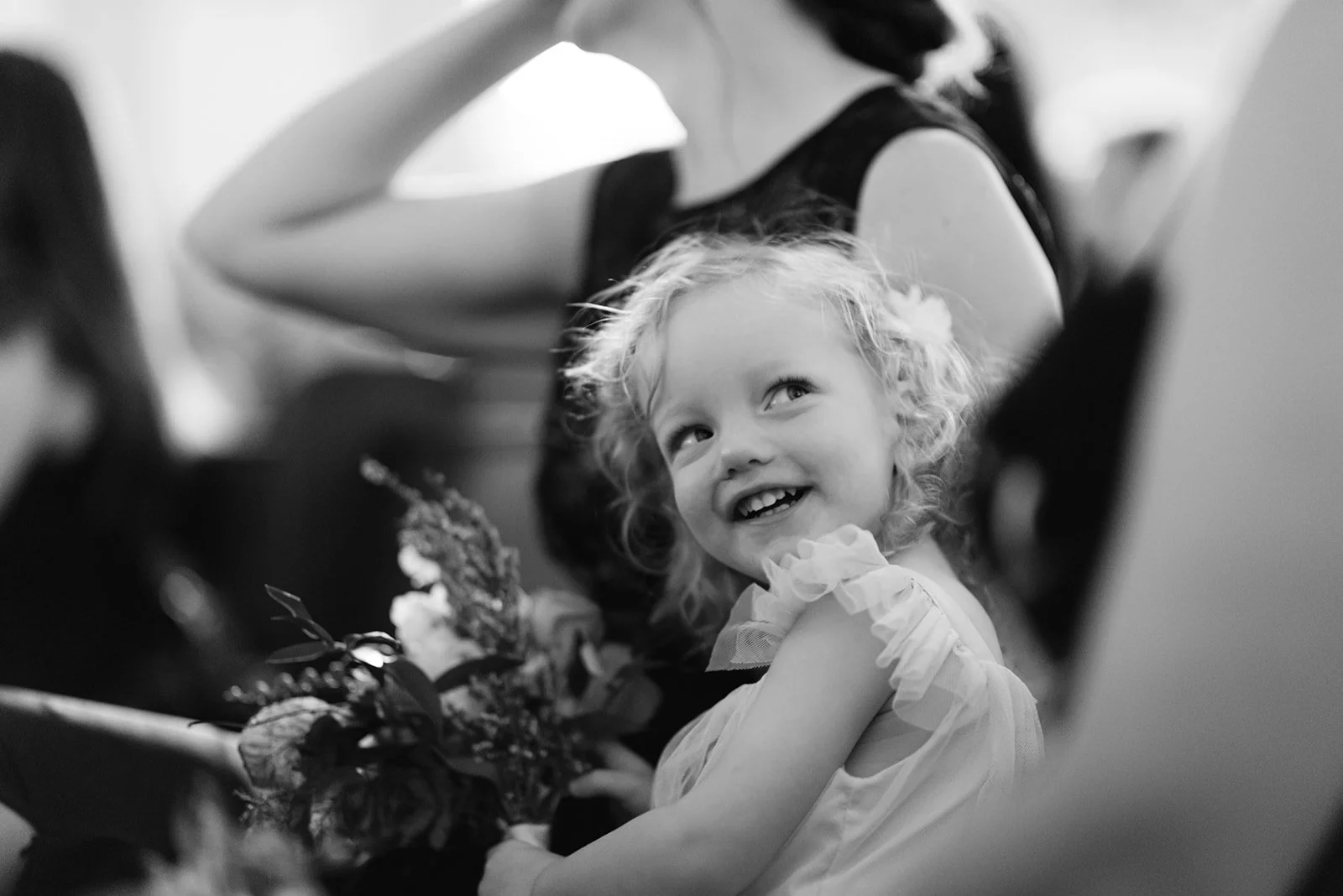 A young girl with curly hair smiling and looking towards the camera, holding a bouquet of flowers, with an adult woman beside her in the background.