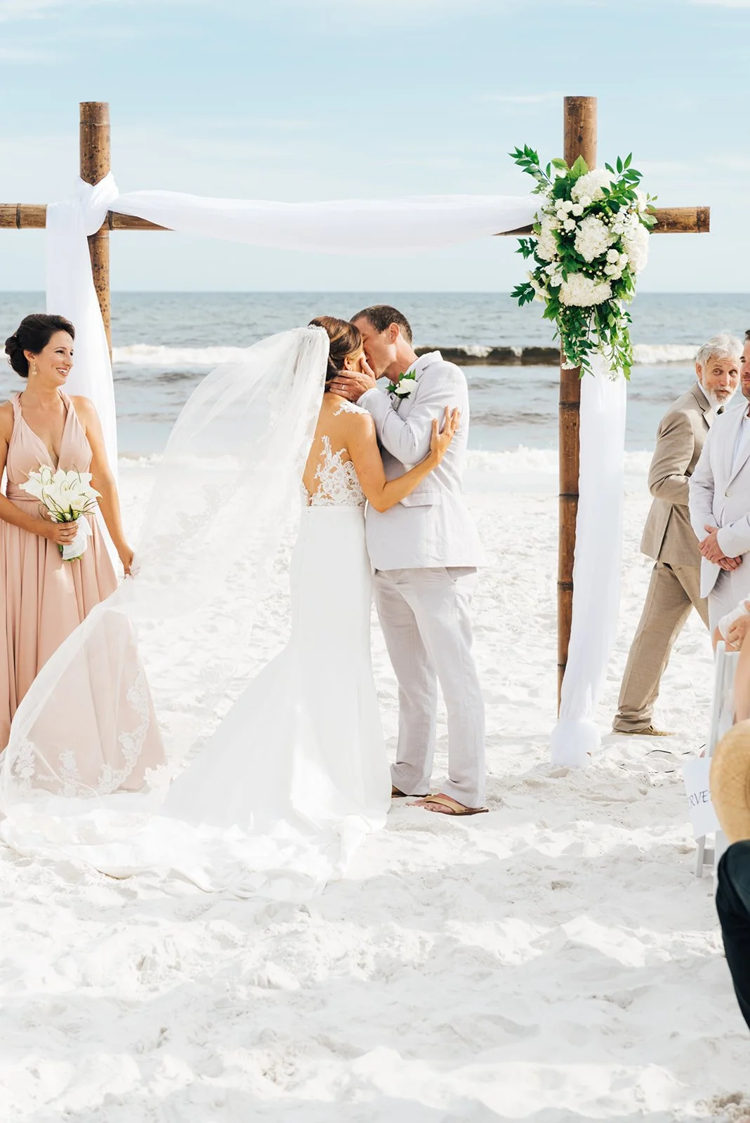 A couple getting married on the beach, kissing under a wooden arch decorated with white flowers and greenery. Bridesmaid holding a bouquet of white flowers, other guests present.