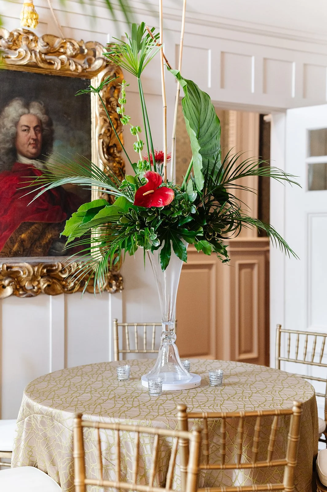 Elegant floral centerpiece with tropical greenery and a red anthurium flower in a tall glass vase on a decorated round table, with gold chairs and a gold-framed portrait in the background.