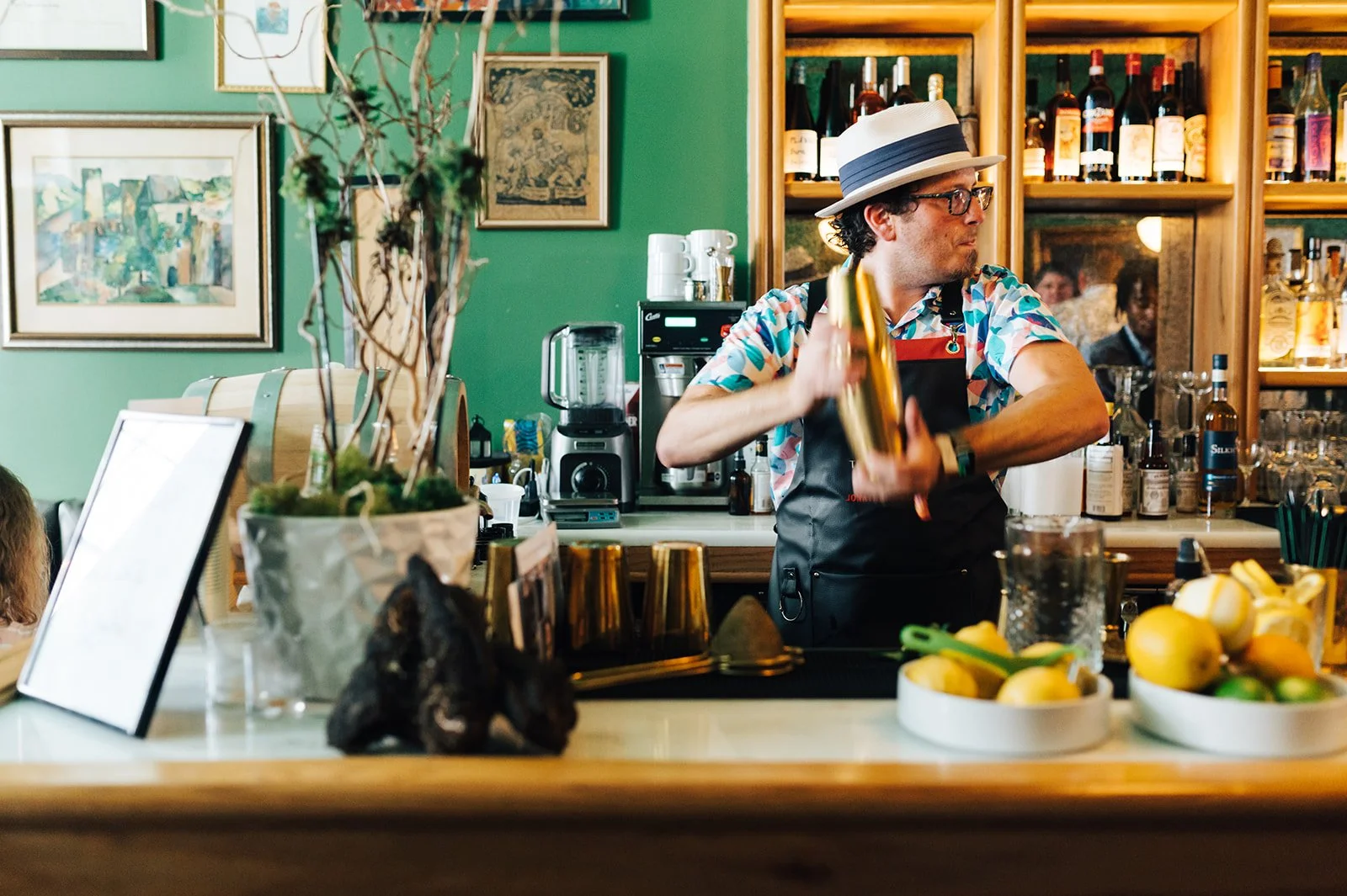 A bartender wearing a hat, glasses, and a colorful shirt with an apron, shakes a cocktail shaker behind the bar. The background includes shelves with liquor bottles, a coffee machine, and framed pictures on a green wall. There are bowls of lemons and