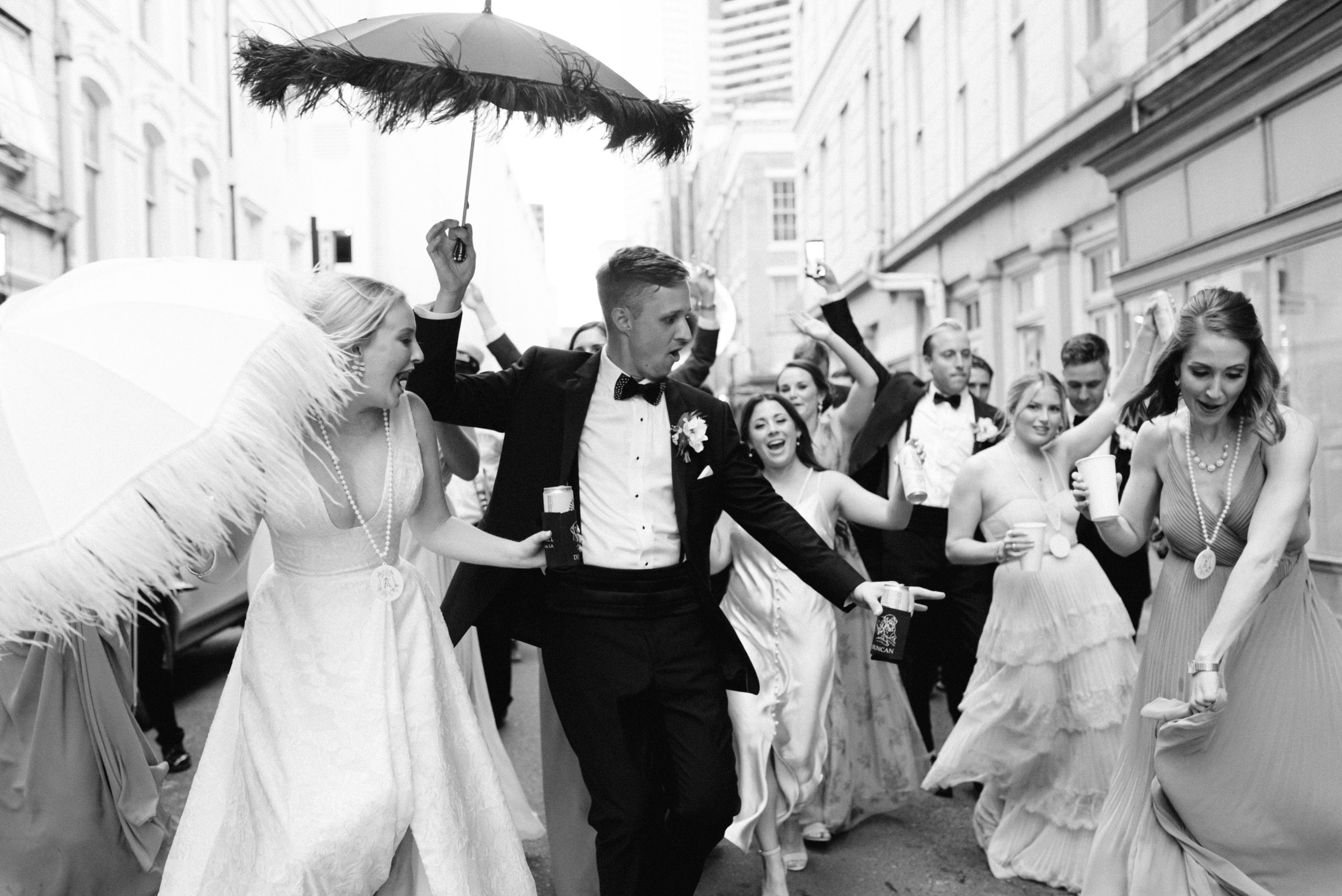 A group of people dressed in formal attire dancing and celebrating on a city street during a wedding or festive event, with some holding drinks and umbrellas.
