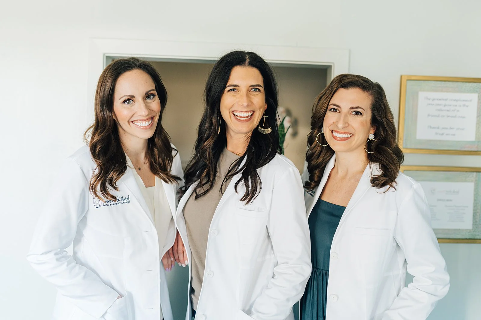 Three smiling women in white medical coats standing together indoors.