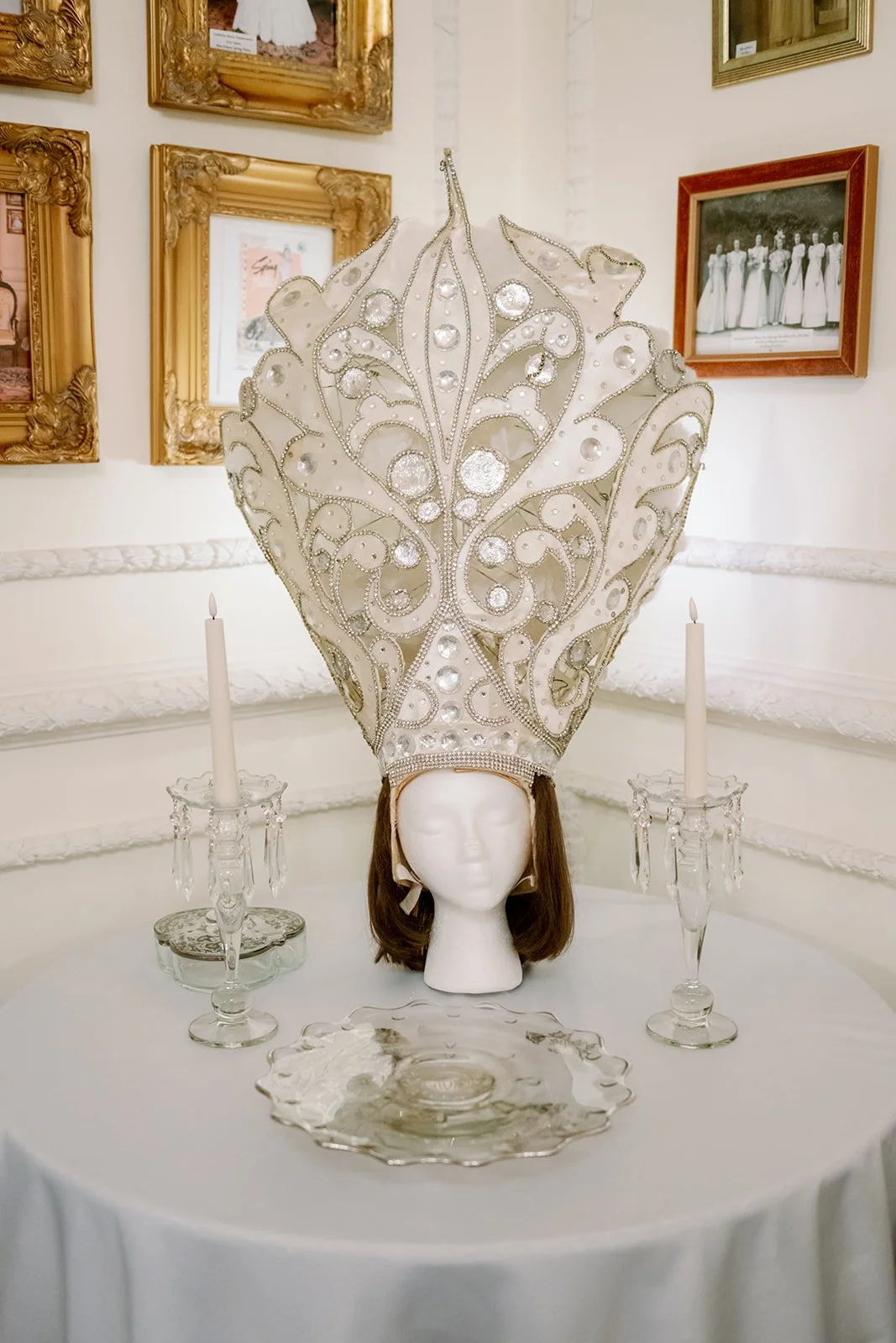 An ornate, large white headpiece with silver embellishments and crystals on a mannequin head, flanked by two glass candleholders with white candles, on a round table in an art gallery setting.