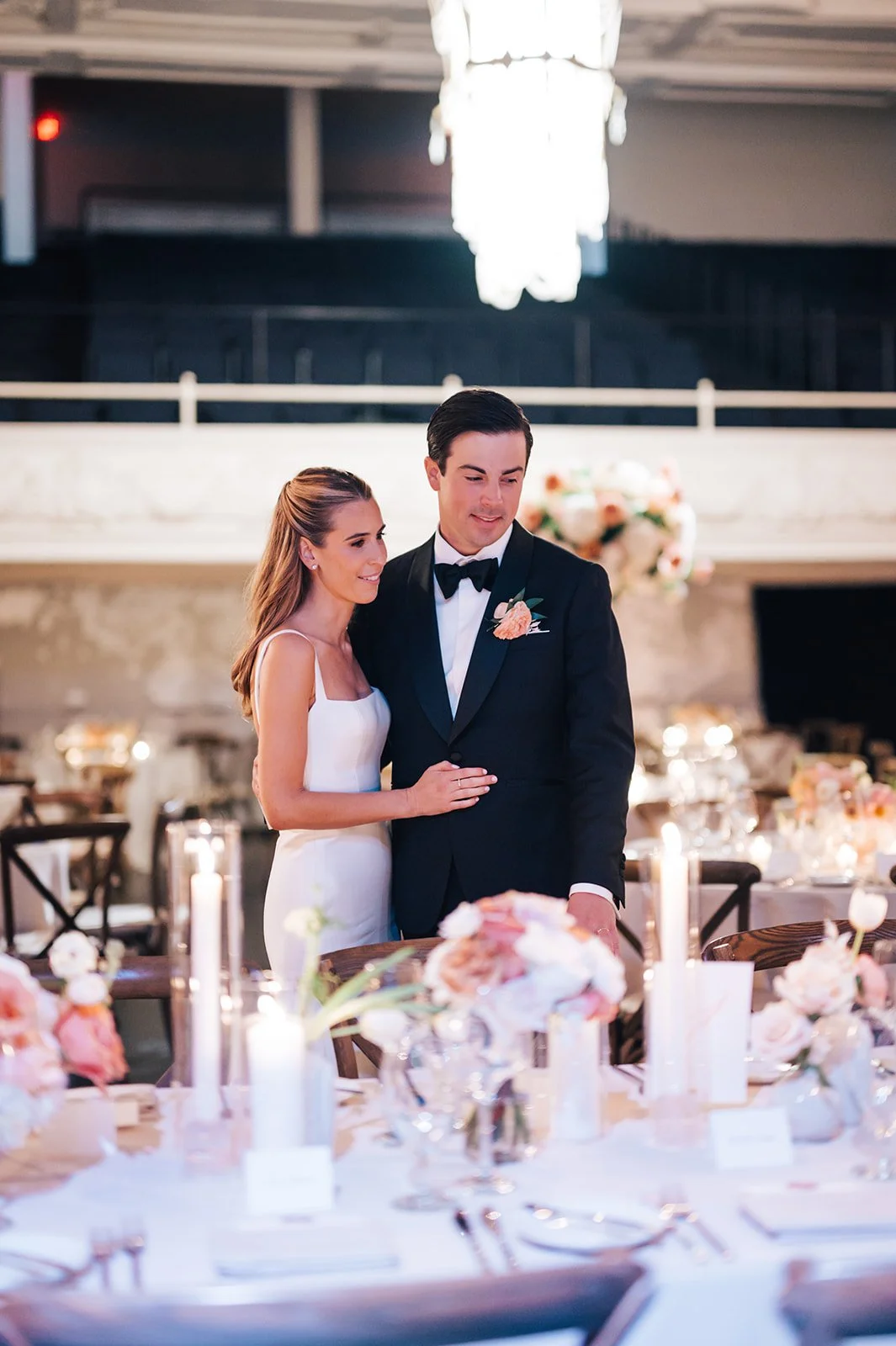 A bride and groom at their wedding reception, standing close together and smiling, surrounded by decorated tables and floral arrangements.