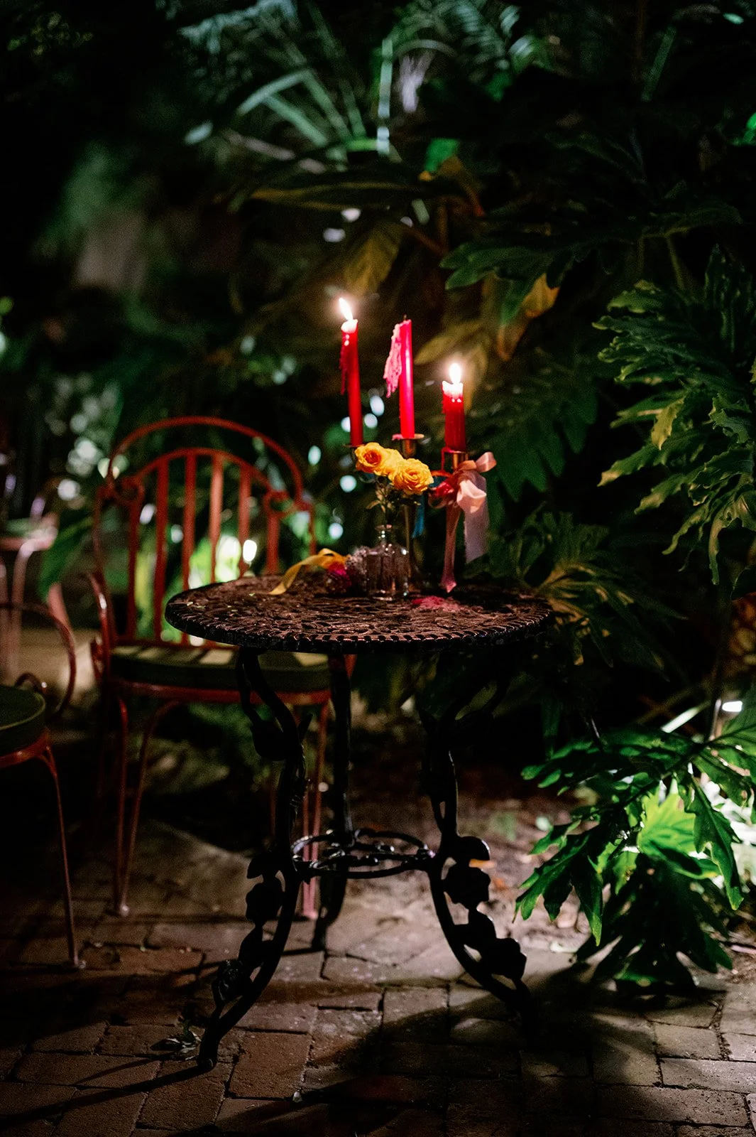 A small round metal table with a decorative design holds a candelabra with three lit red candles, surrounded by yellow flowers, set in a lush garden at night with green foliage and a brick pathway.