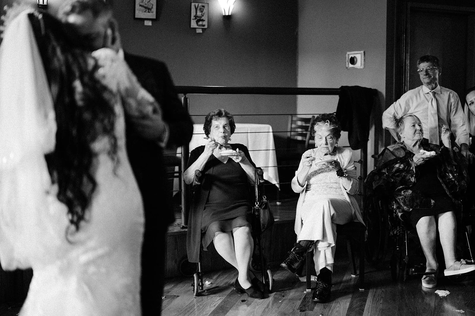 Black and white photo of elderly women dining at a social event, with three women seated in the background, and two people standing in the foreground, partially visible.