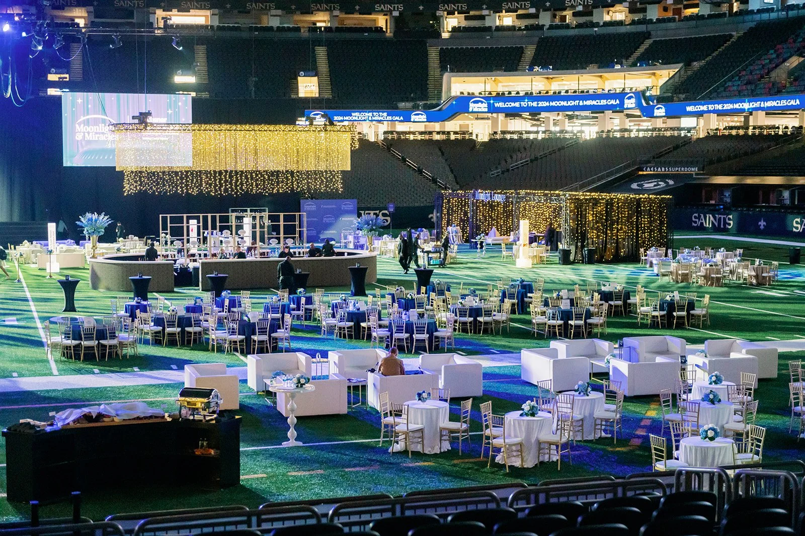 Event setup in a stadium with round and rectangular tables, white sofas, and gold chairs, decorated with blue and white flowers, illuminated by string lights and stage lighting for a formal gathering.