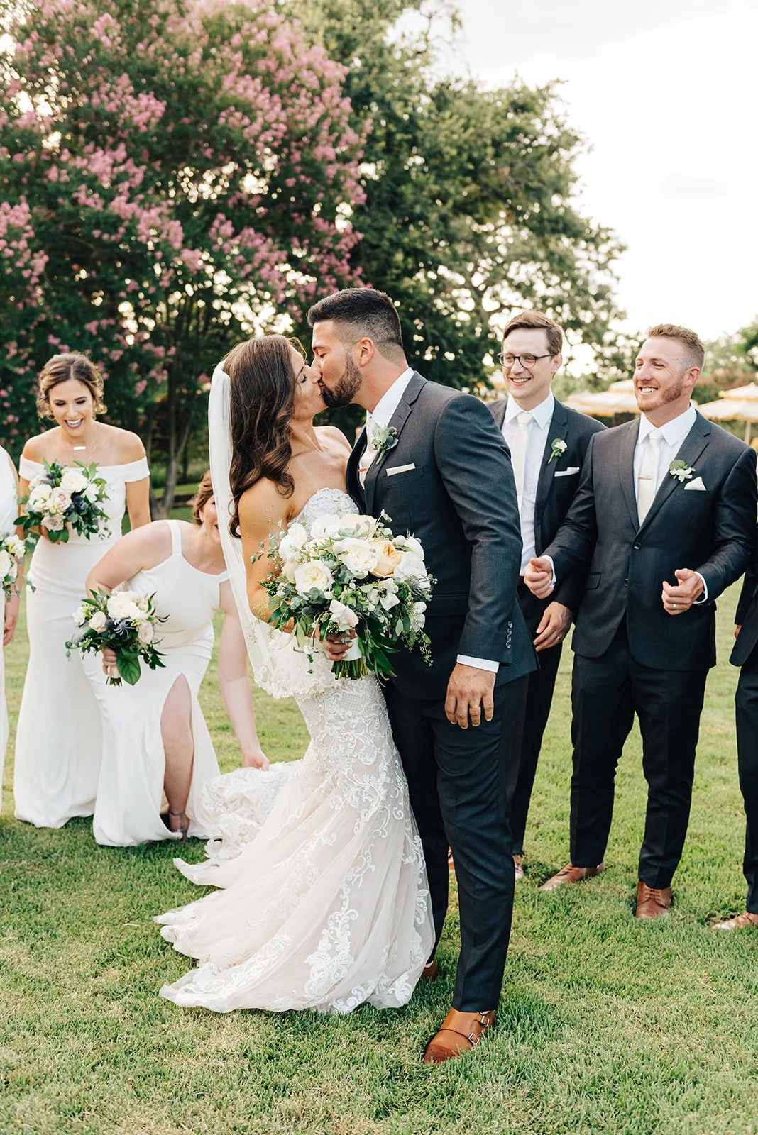 A wedding ceremony outdoors with a bride and groom sharing a kiss, surrounded by bridesmaids and groomsmen. The bride is in a white lace wedding dress and the groom in a dark suit. The bridesmaids wear white dresses, and the groomsmen are in dark suits. The scene is set against trees with pink blossoms, during daylight.
